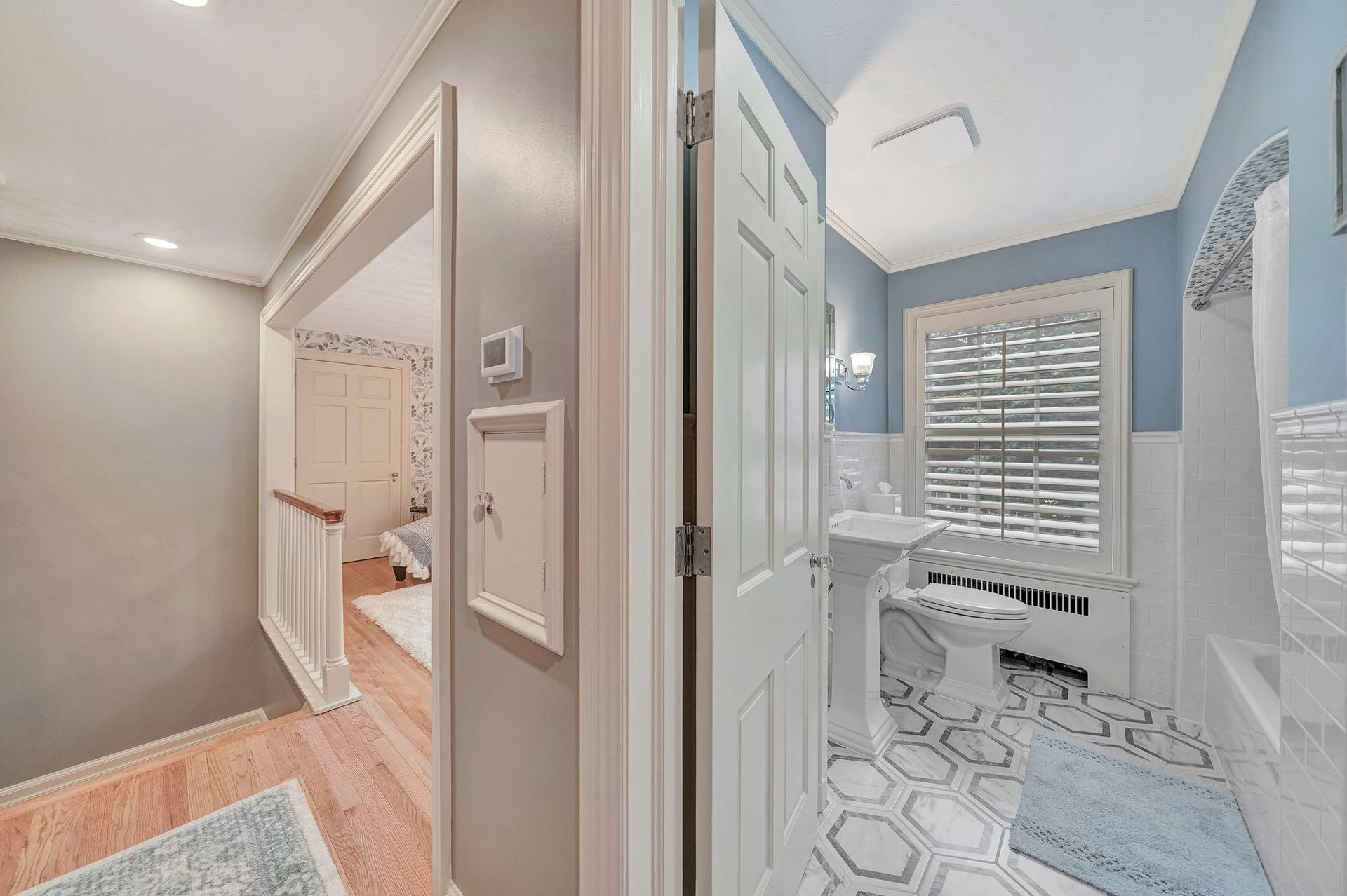 Hallway with view of a bathroom. Light blue and white bathroom with white pedestal sink, toilet, and shuttered window.