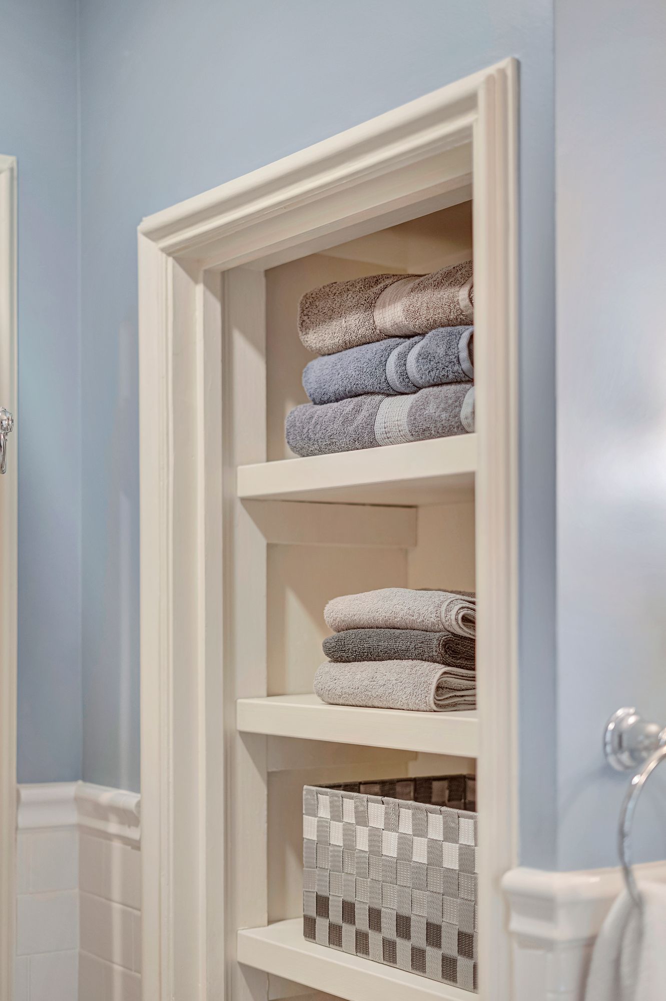 Built-in white shelf in a blue bathroom holding folded towels and a patterned box.