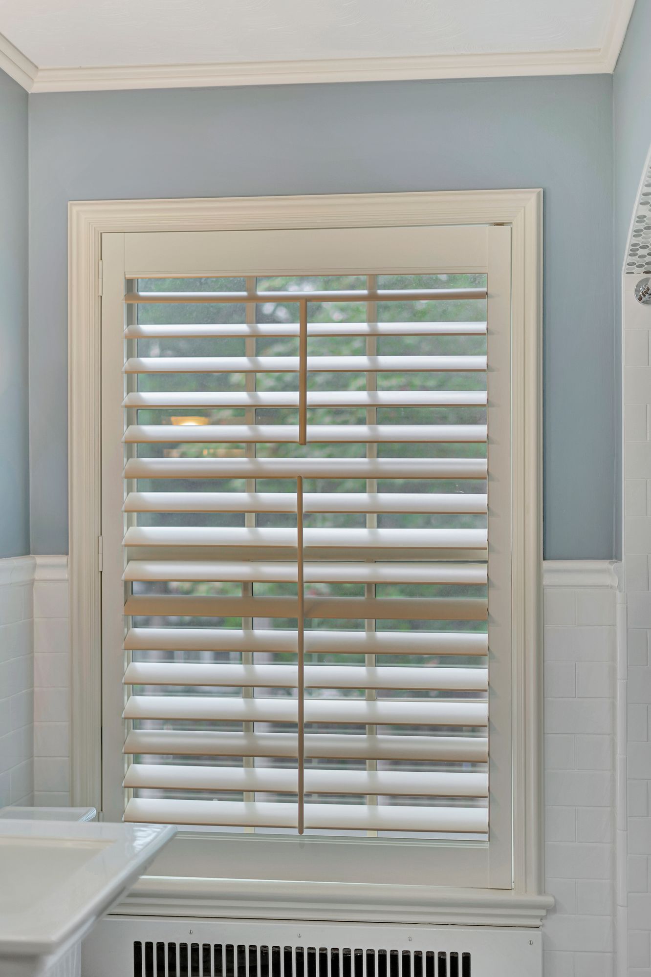 Window with white shutters, light blue walls, and white trim.  Part of a bathroom.