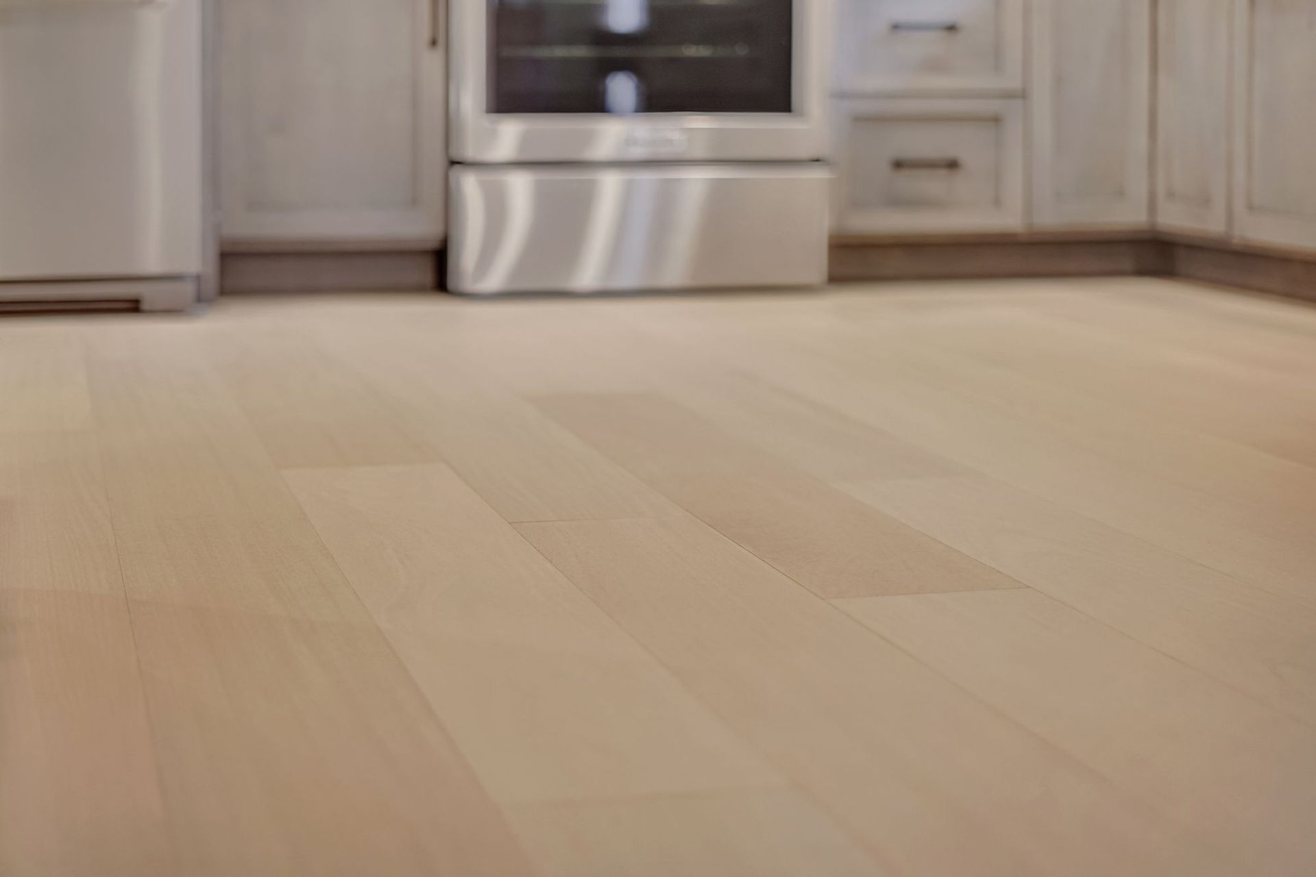 Light-colored wooden floor in a kitchen, with stainless steel appliances and off-white cabinets in the background.