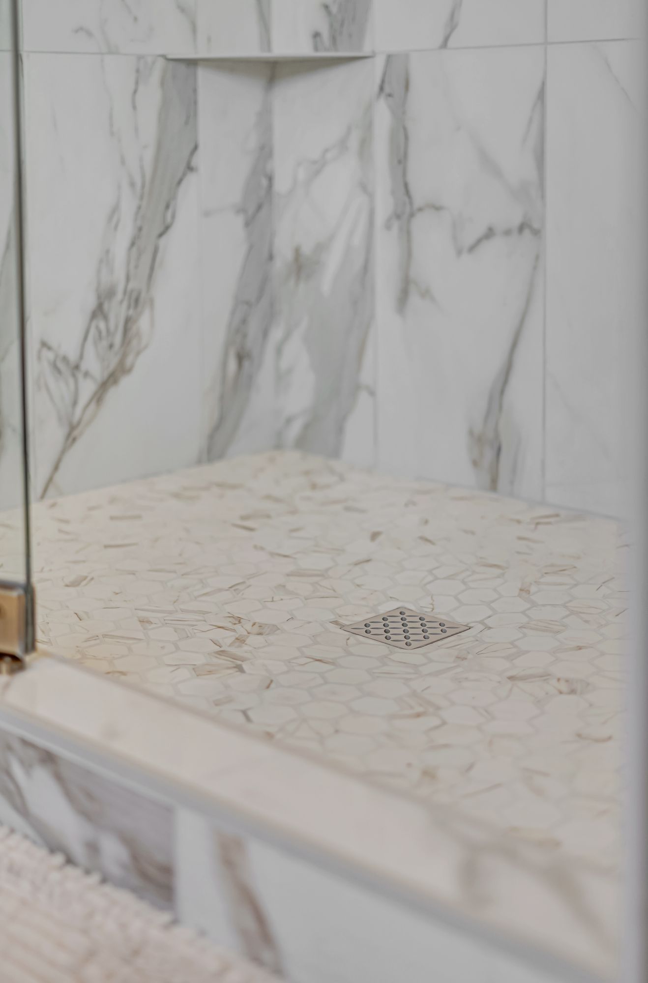 Shower floor with marble-like tile, a drain, and a shelf. Glass shower door is visible.