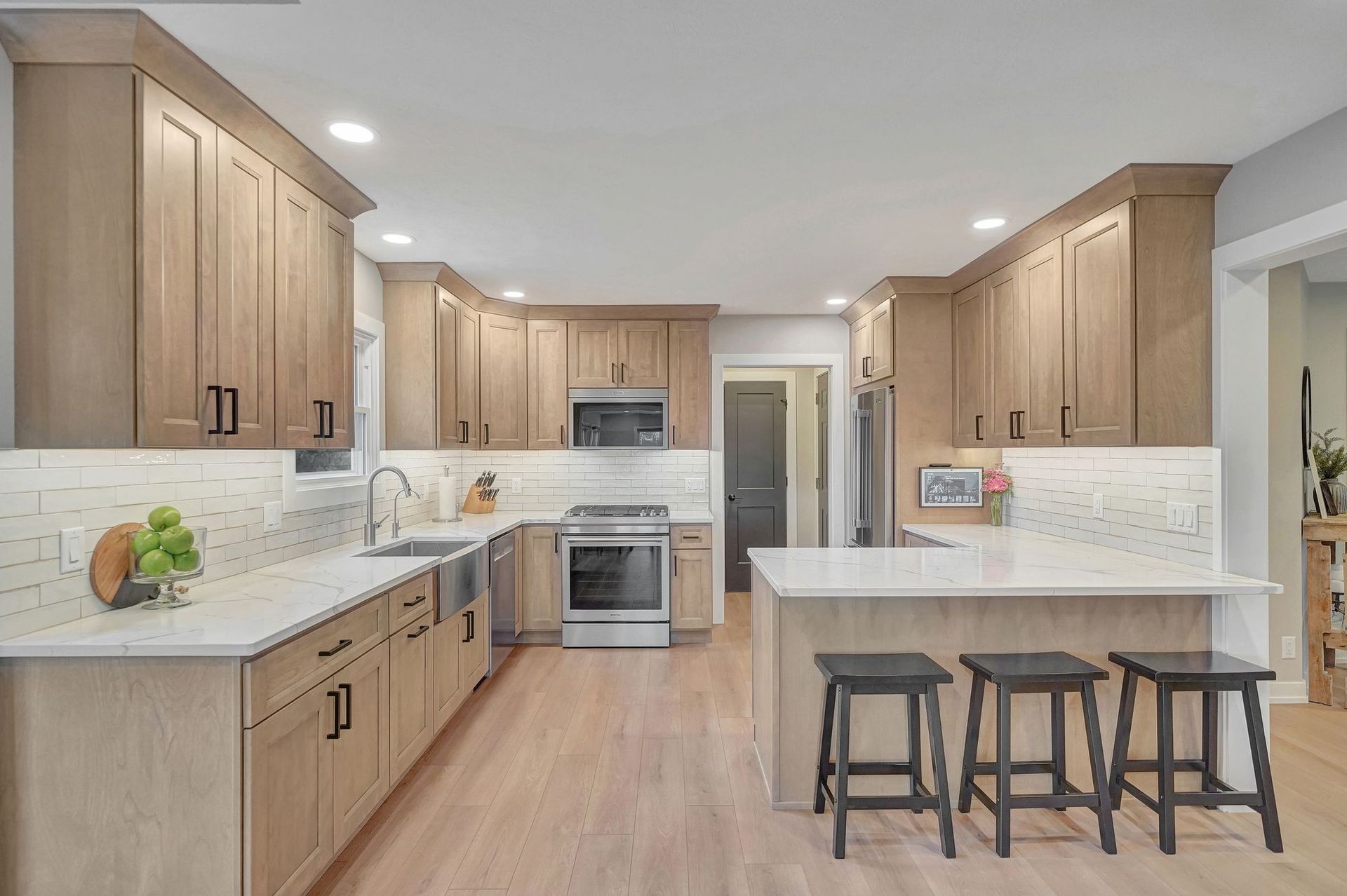 Modern kitchen with light wood cabinets, white countertops, stainless steel appliances, and a breakfast bar with stools.