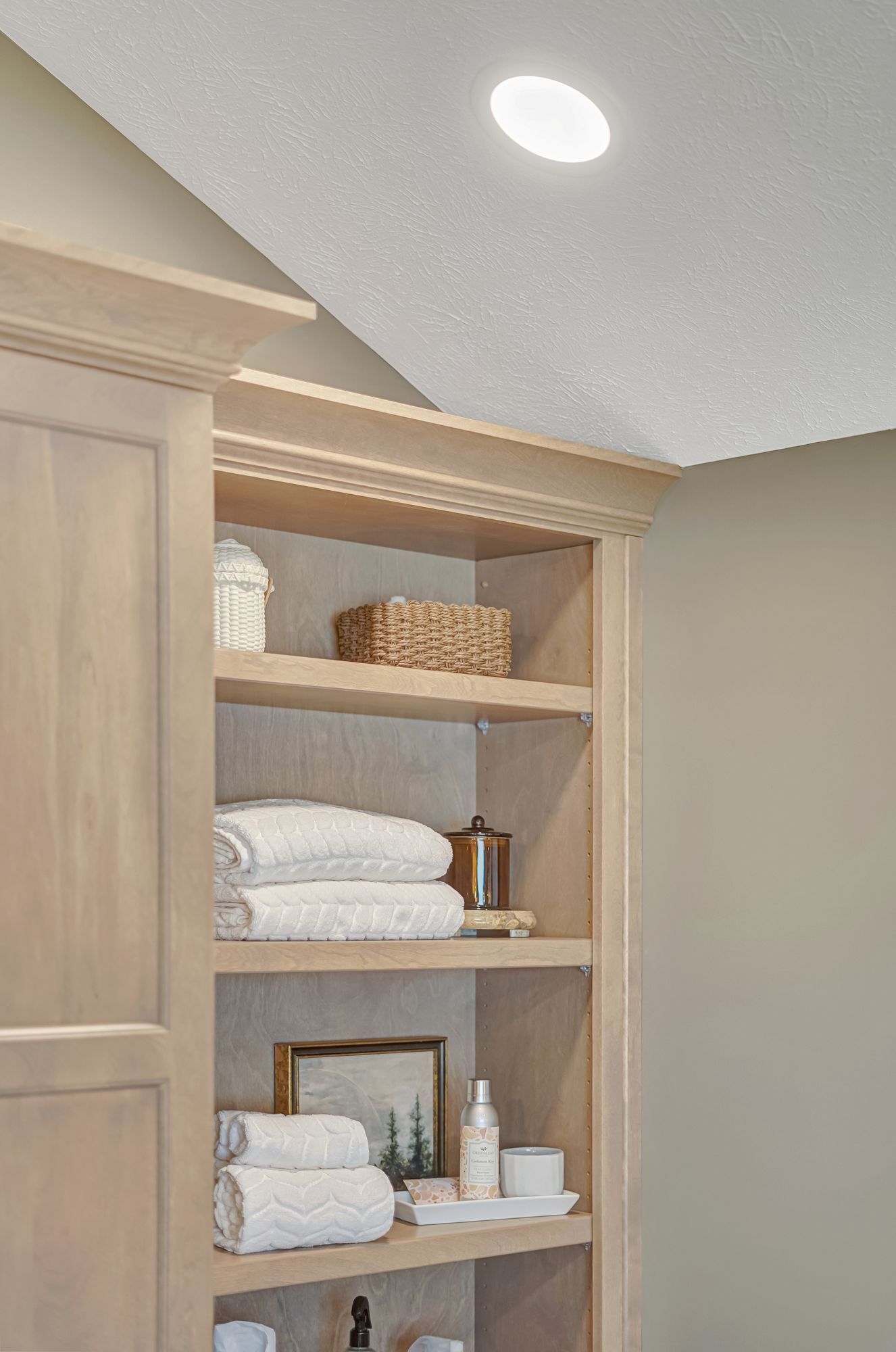 Built-in beige shelving with stacked white towels, a woven basket, and other bathroom items; a recessed ceiling light.