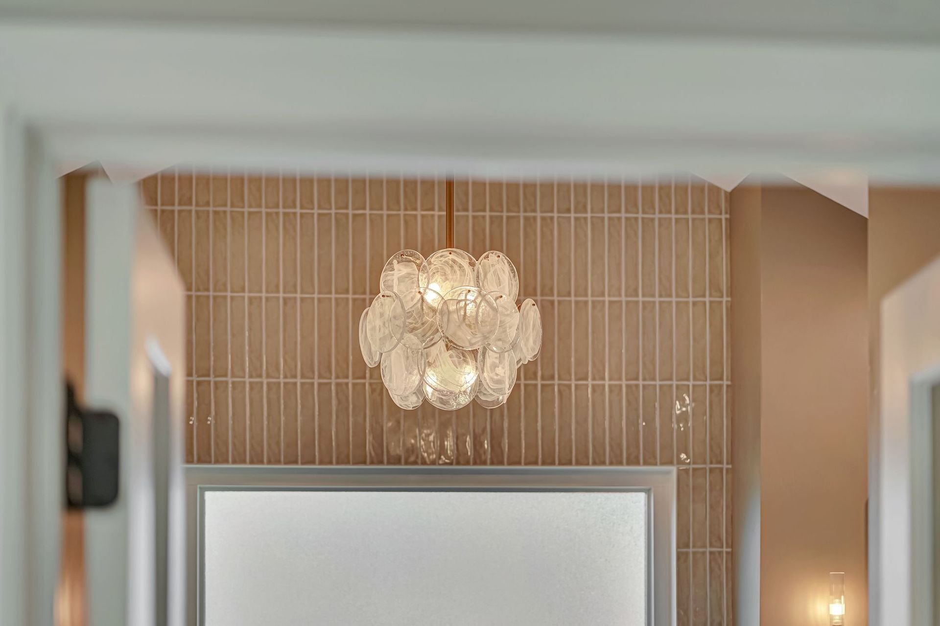 Crystal chandelier in bathroom with tan tiled wall, viewed through a doorway.