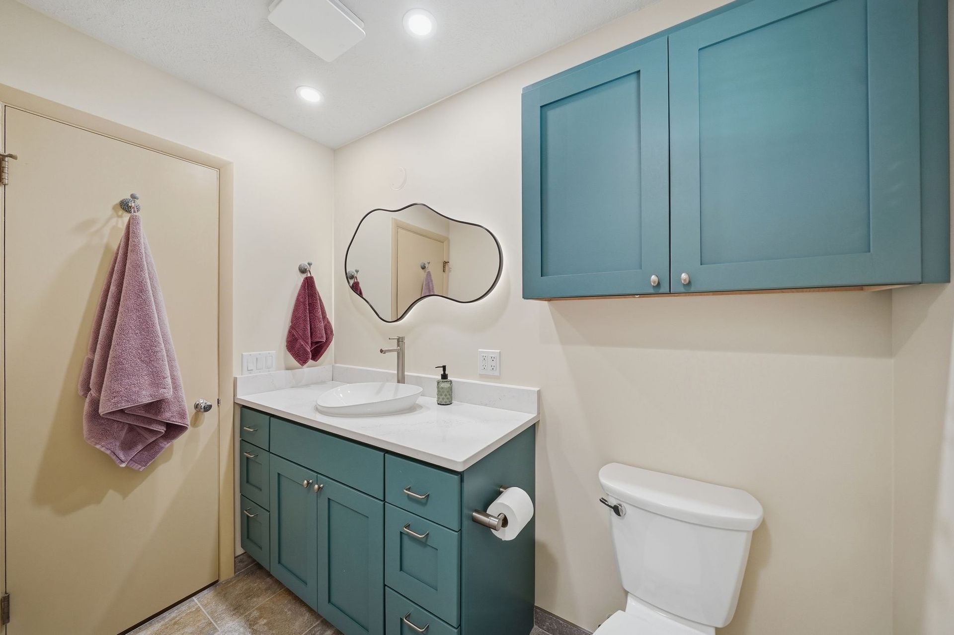 Compact bathroom with teal cabinets, white sink, wall mirror, towel hooks, and toilet.