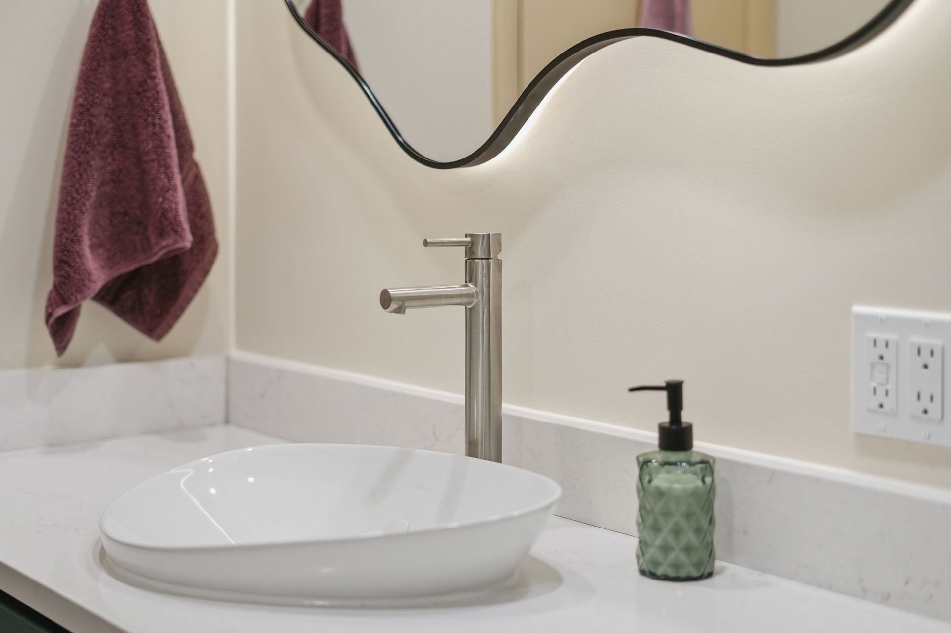 Modern bathroom vanity with white sink, brushed metal faucet, and green soap dispenser below wavy mirror