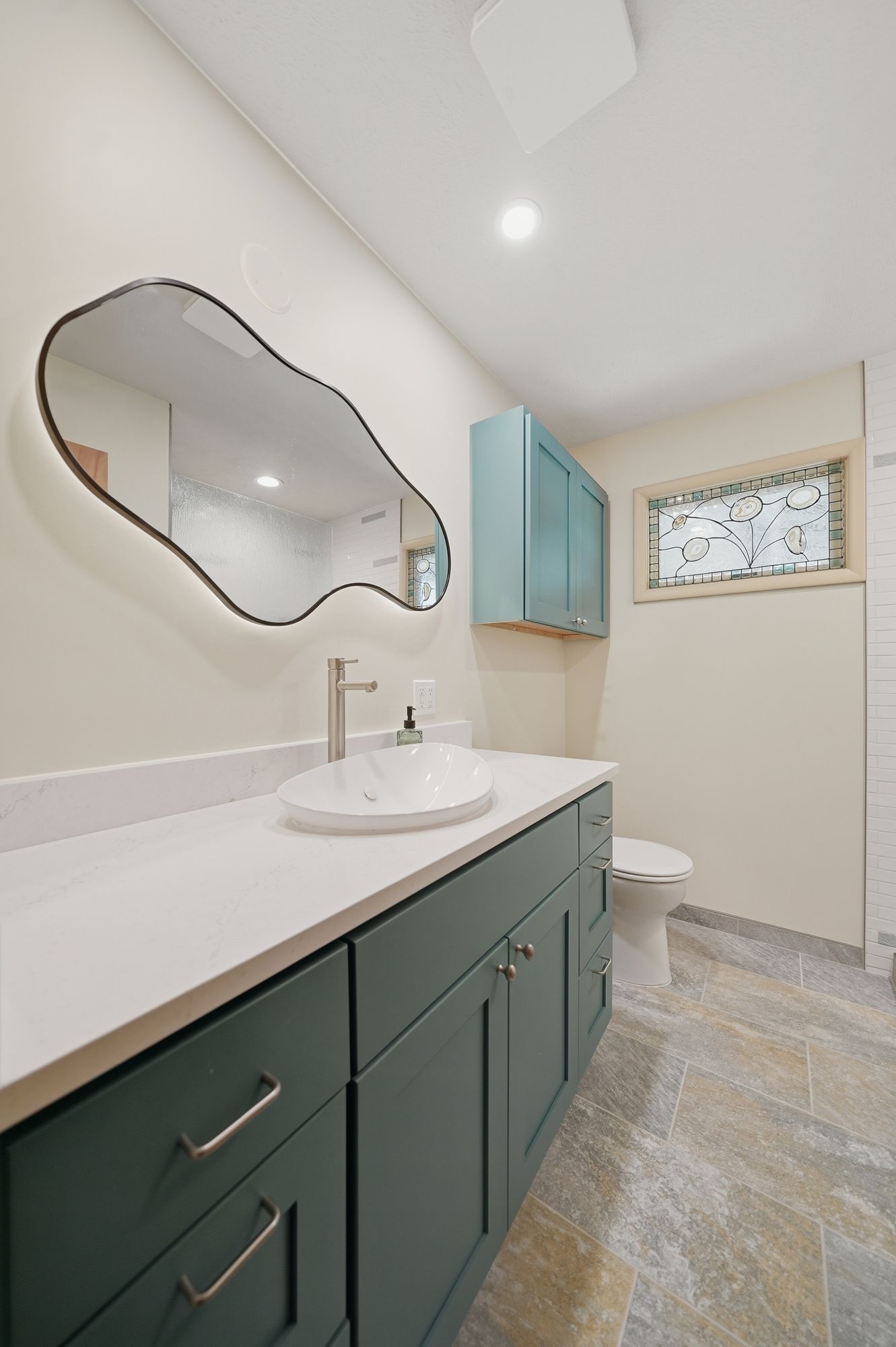 Small bathroom vanity with green cabinets, white sink, wavy mirror, and frosted window.