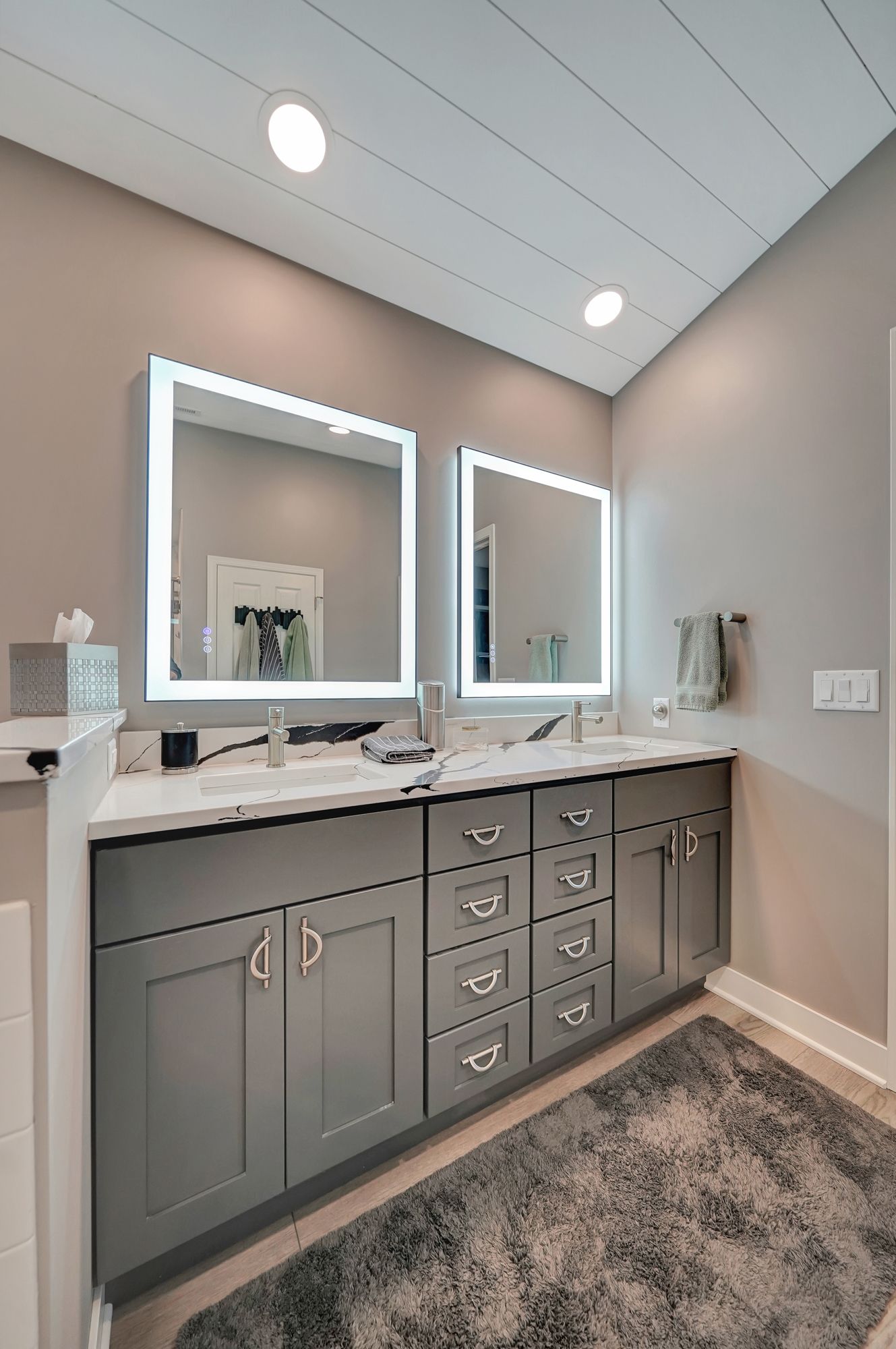 Modern bathroom with gray vanity, two backlit mirrors, white countertops, and gray rug.