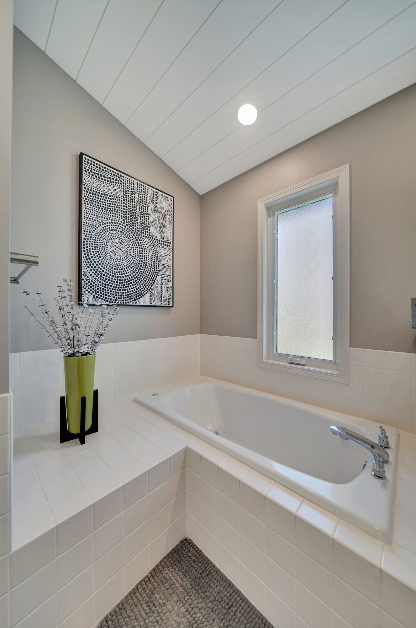 Bathroom with white tub, white tile, and grey walls. Artwork and window present.