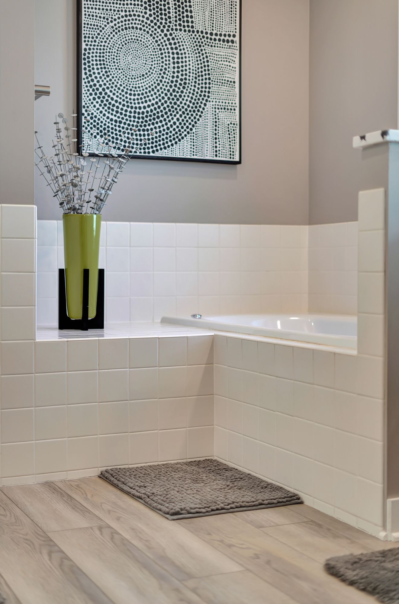 Modern bathroom with white tiled walls, a bathtub, and grey rug. A green vase with flowers sits in a niche.