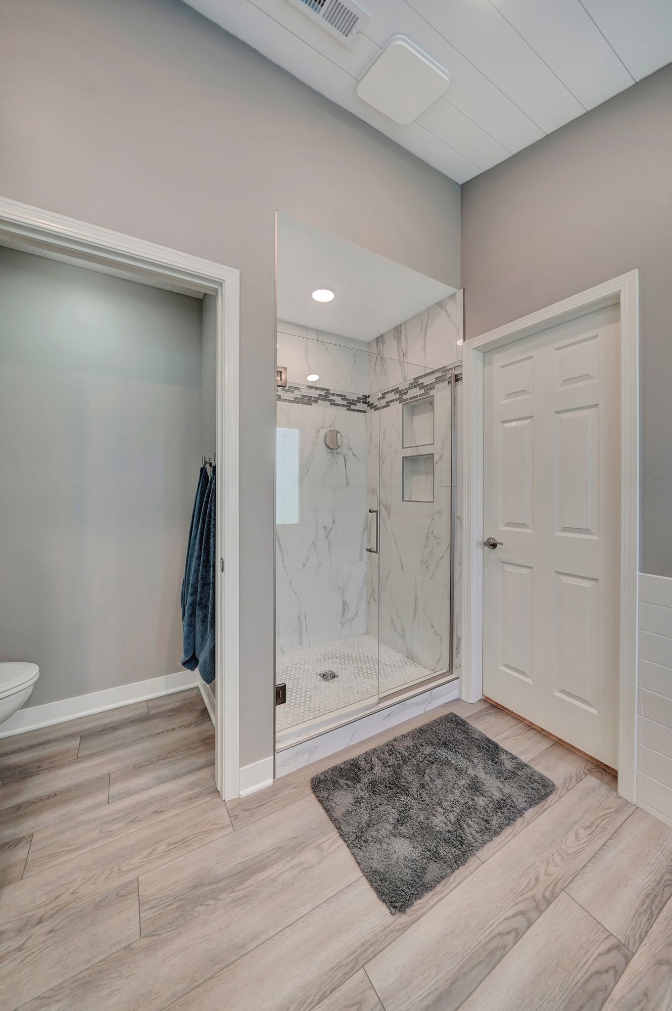 Bathroom with shower and door; gray walls, wood-look floor, white trim, shower has marble-look tile.