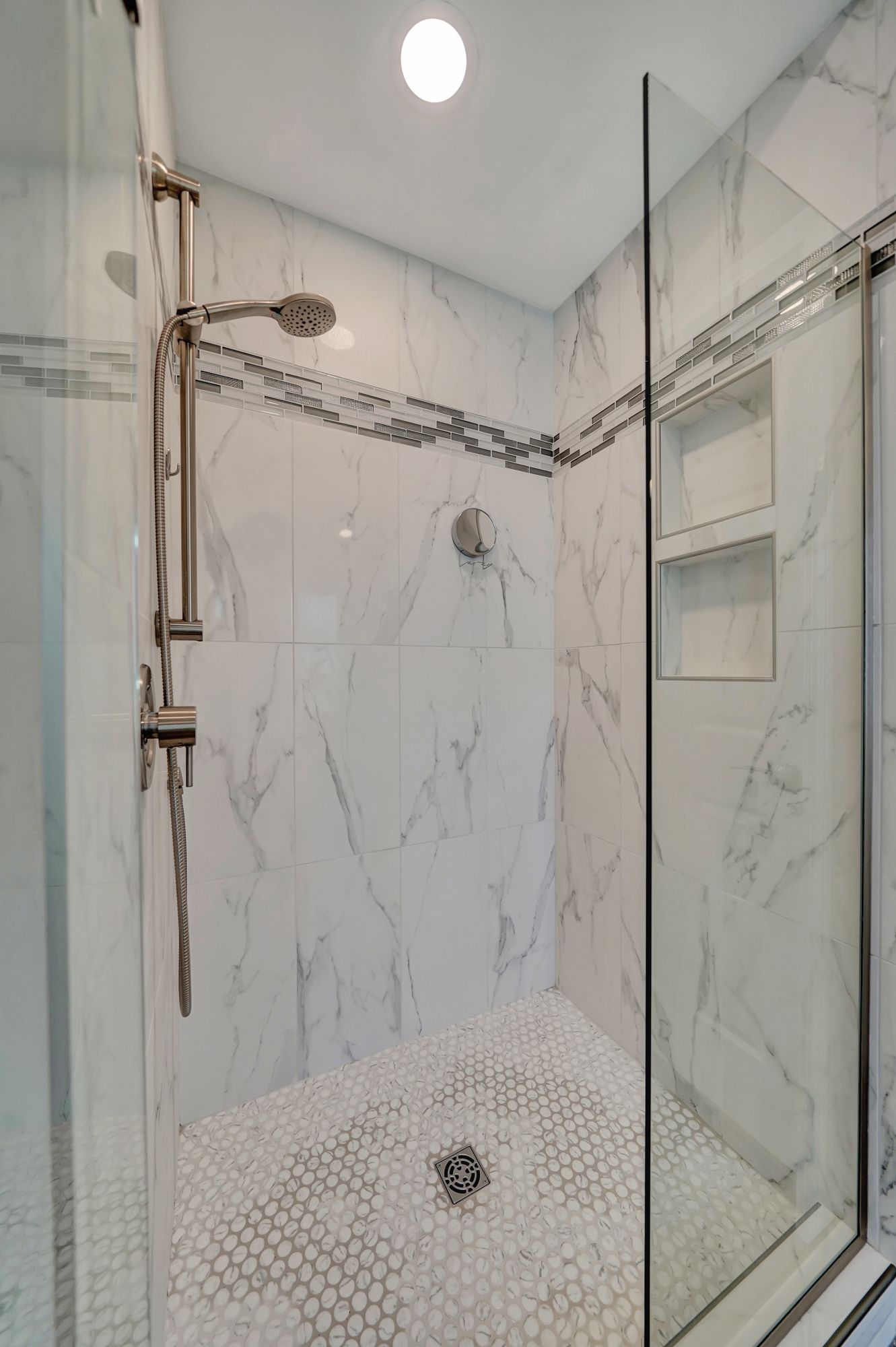 Tiled shower with glass door, marble walls, and pebble floor. Built-in shelves and silver shower fixtures.
