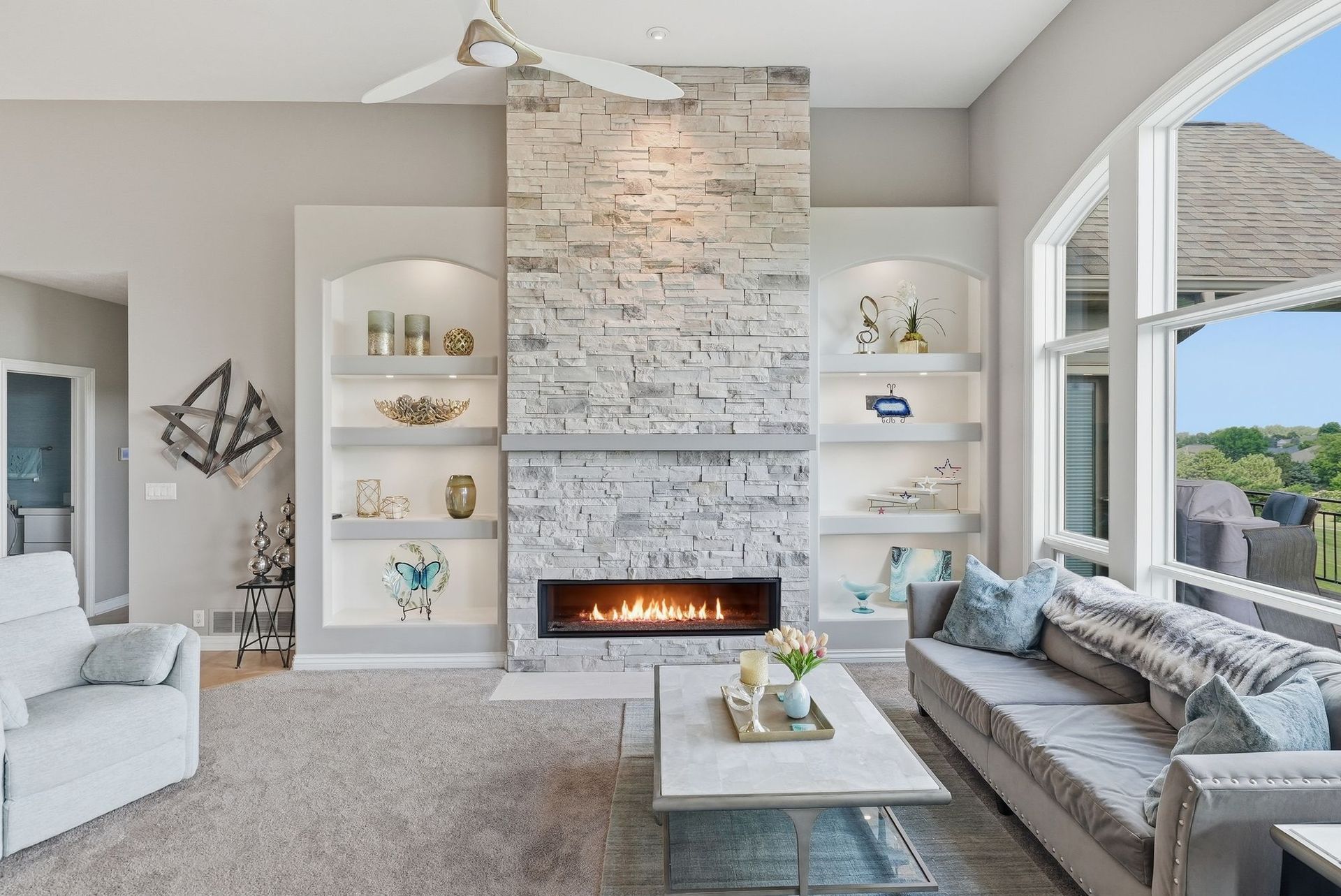 Bright modern living room with stone fireplace, built-in shelves, and large windows overlooking greenery