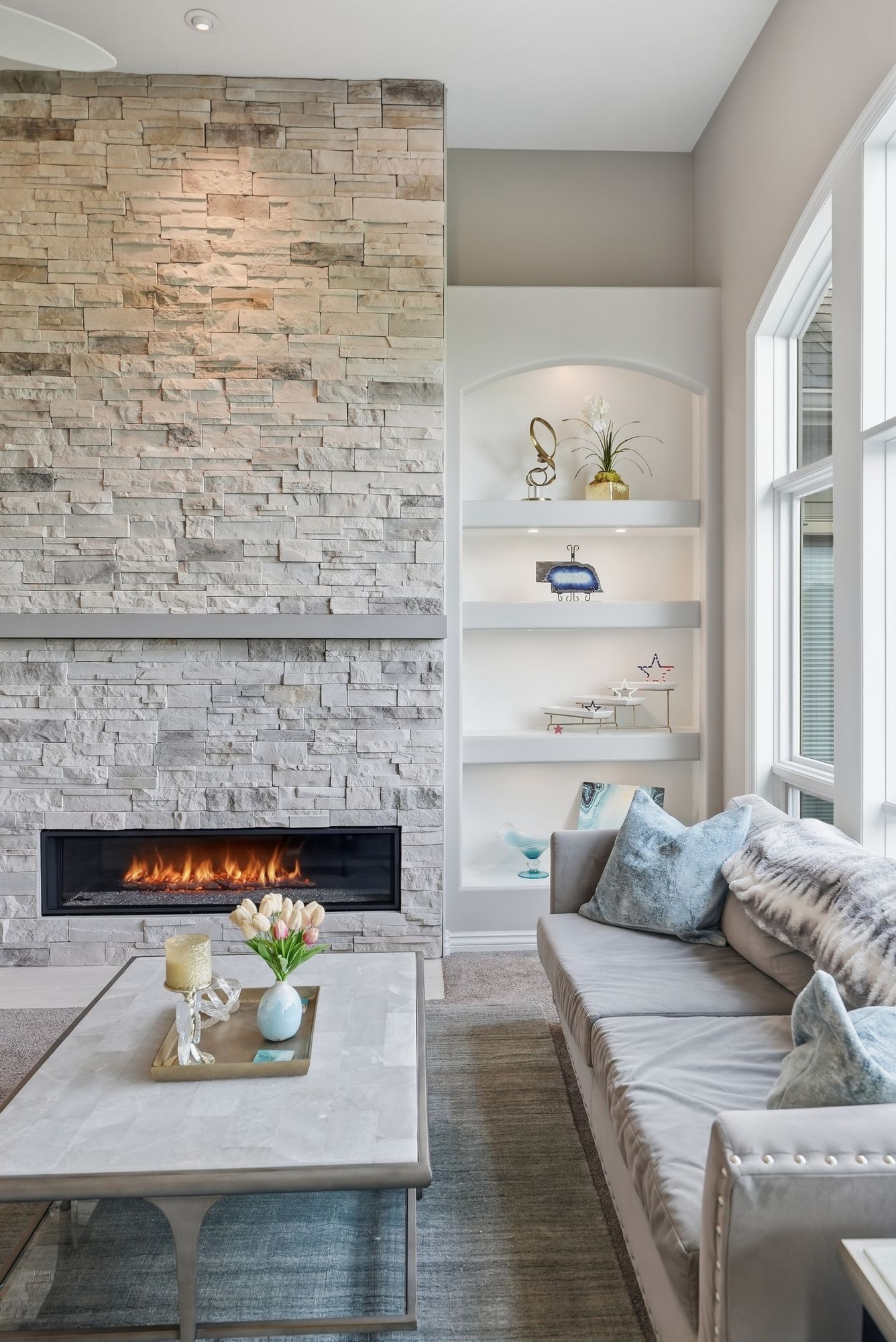 Modern living room with stone fireplace, built-in shelves, and a gray sofa by a bright window.