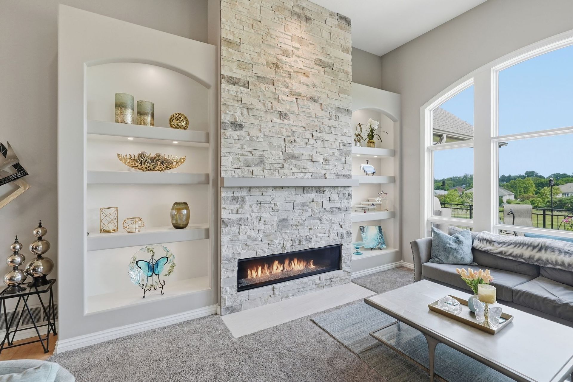Bright living room with stone fireplace, built-in shelves, and large windows overlooking water