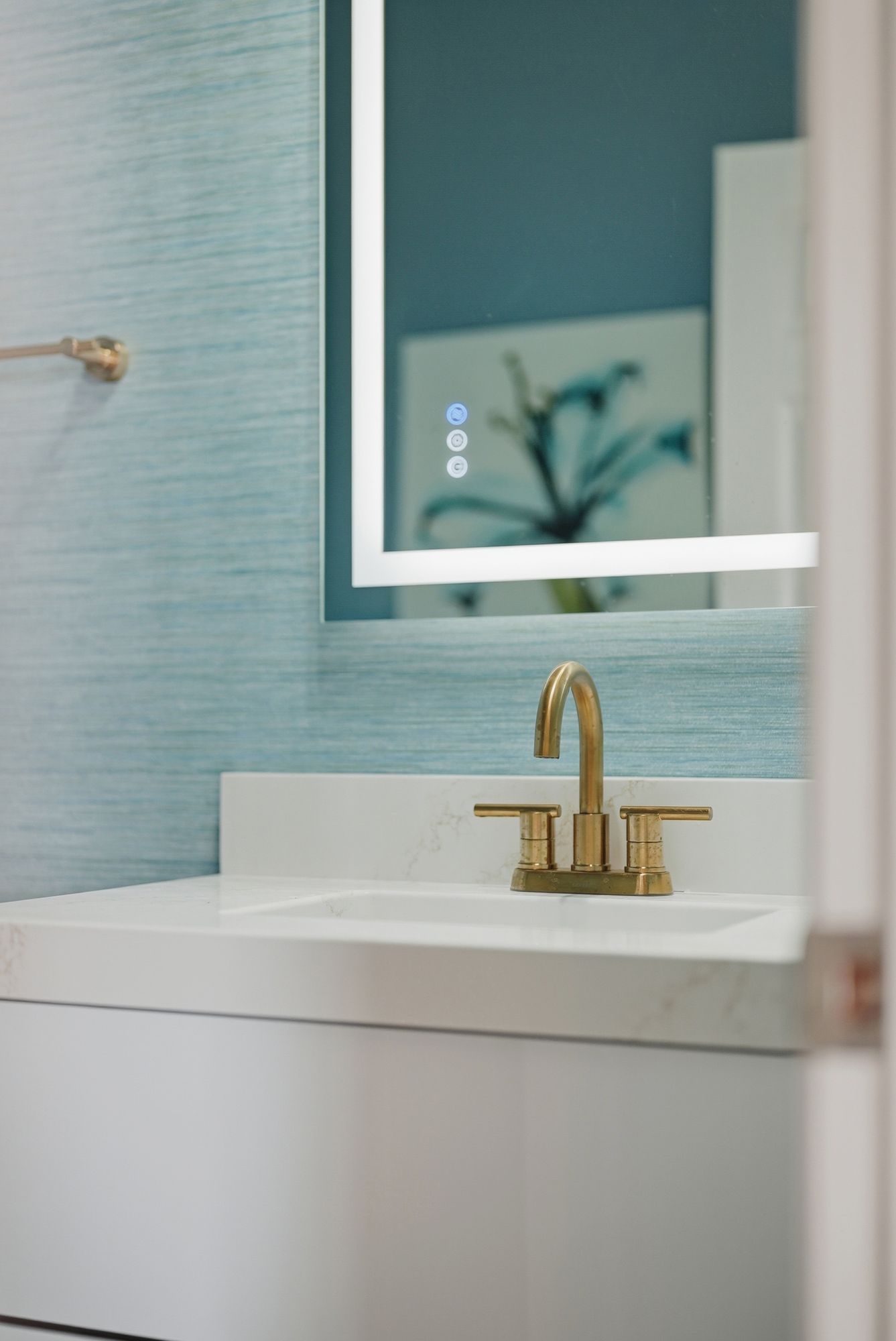 White bathroom vanity with brass faucet and lit mirror against blue textured wall
