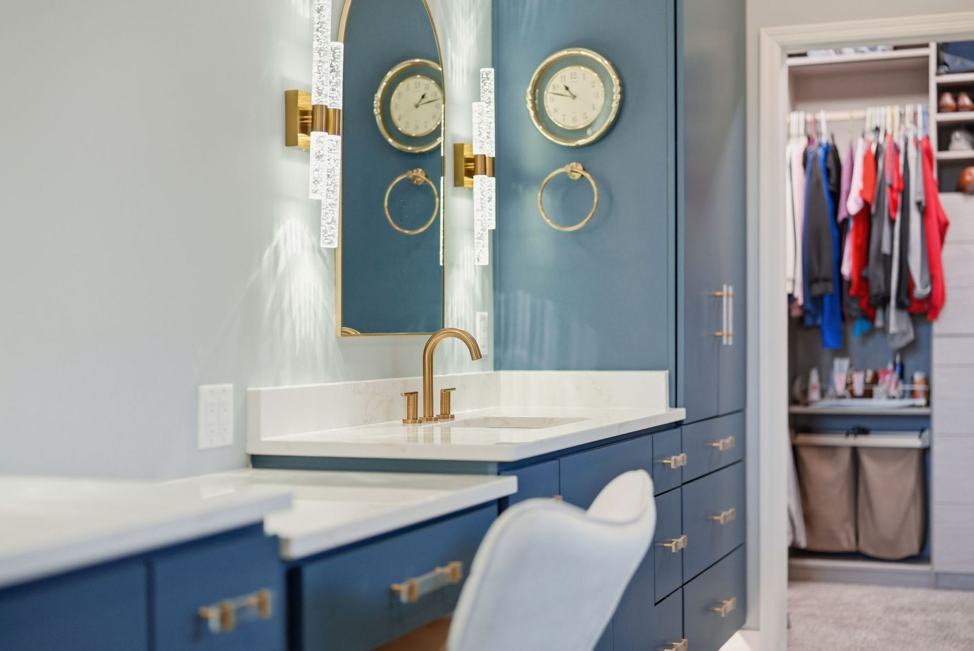Blue bathroom vanity with gold fixtures, round mirrors, and an open closet beside it