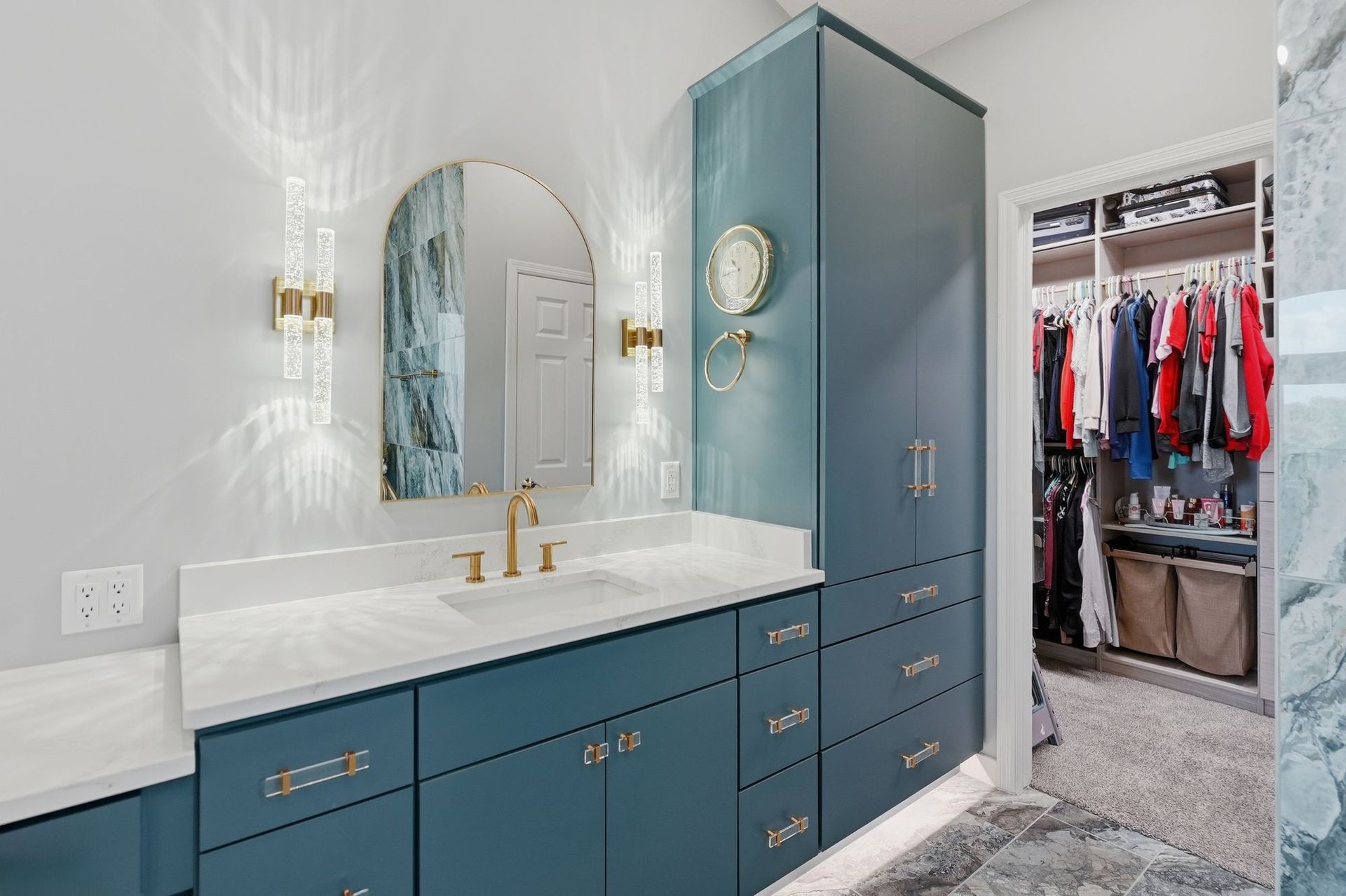 Blue bathroom vanity with marble countertop, gold fixtures, arched mirror, and open closet doorway