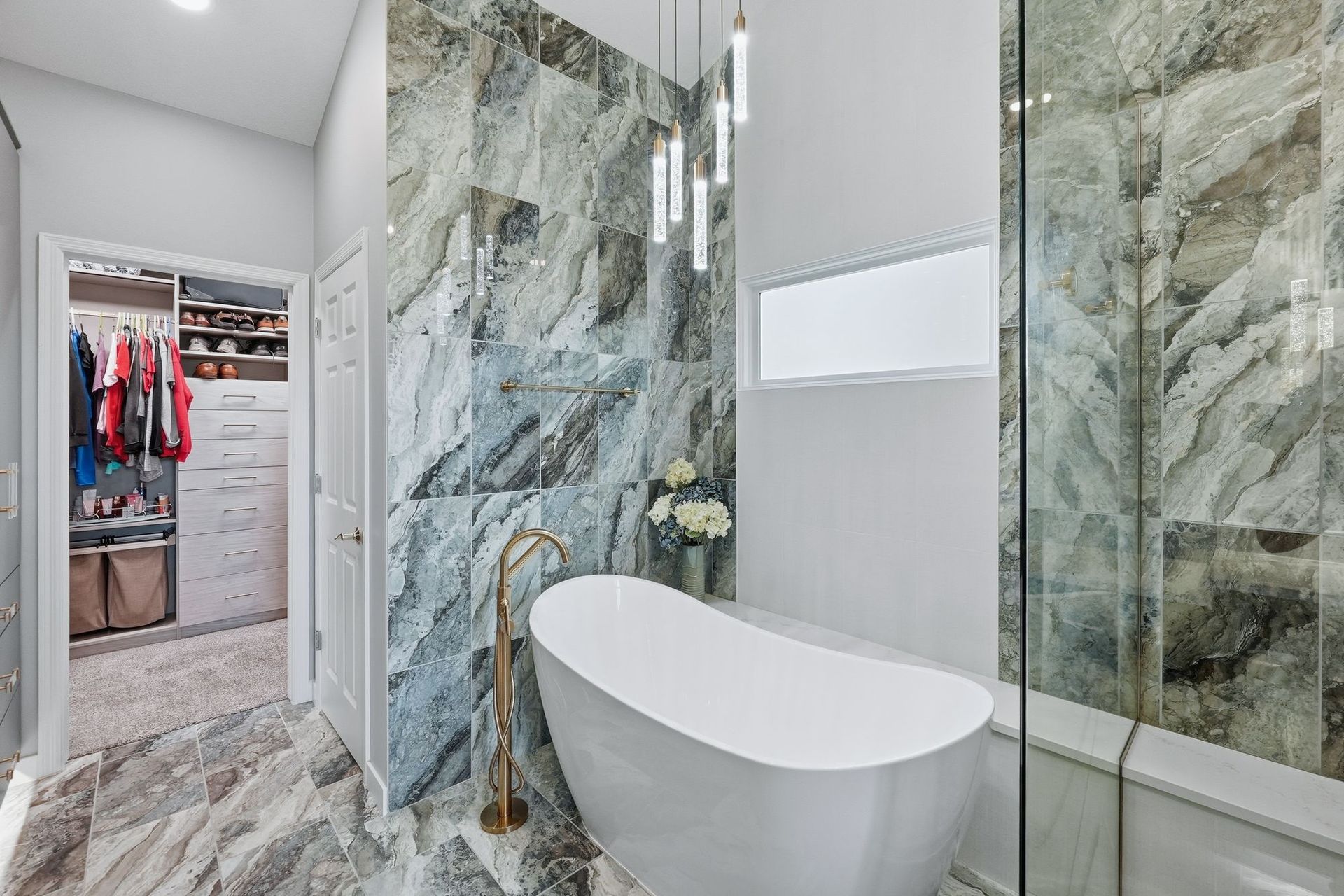 Modern bathroom with marble walls, glass shower, freestanding tub, and open closet visible through the doorway