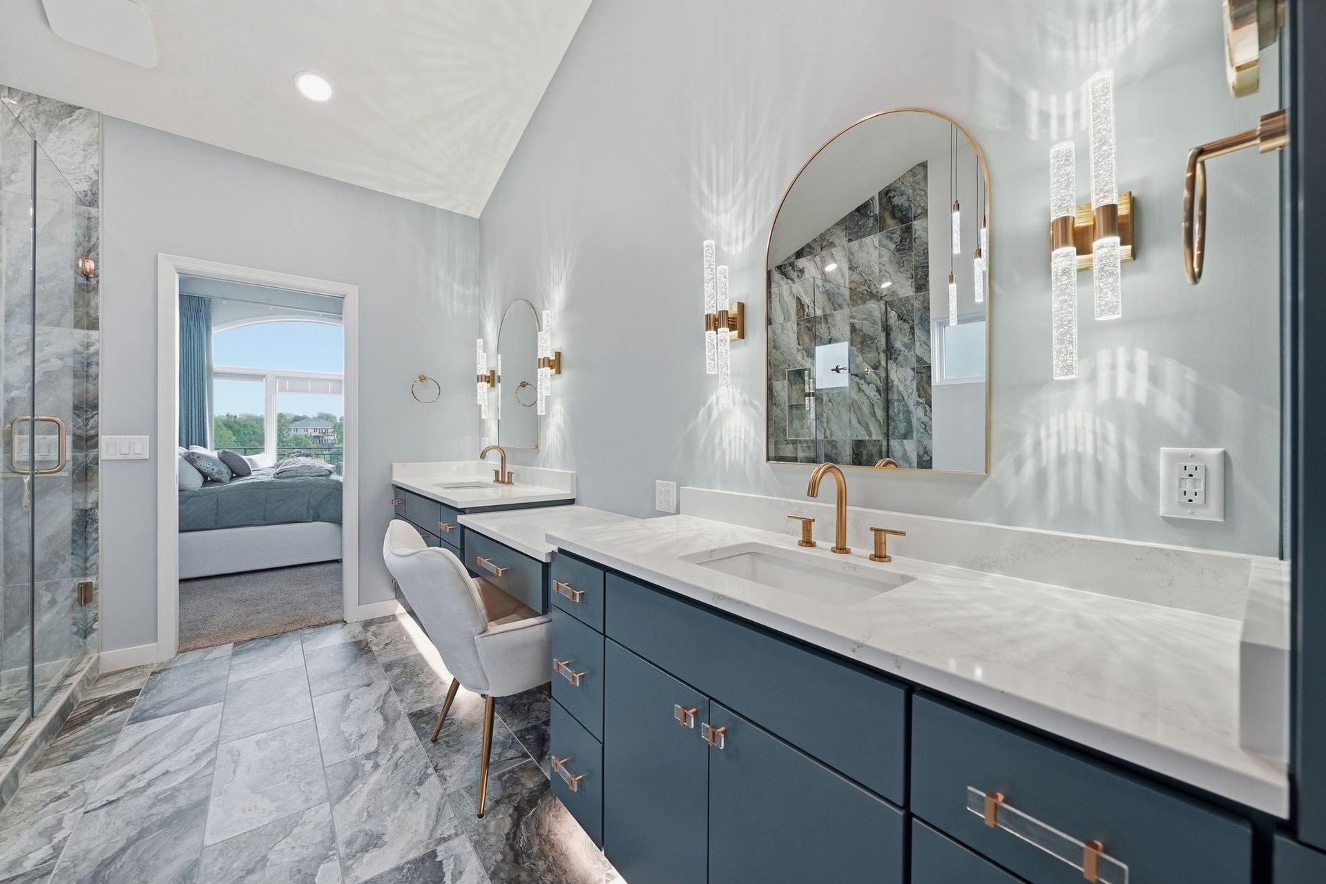 Bright modern bathroom with blue vanity, marble countertop, gold fixtures, and arched mirror.