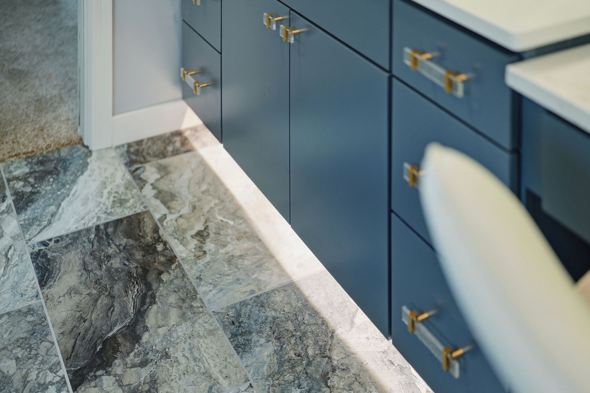 Blue bathroom vanity with brass handles and marble floor tiles, with a white chair in the foreground