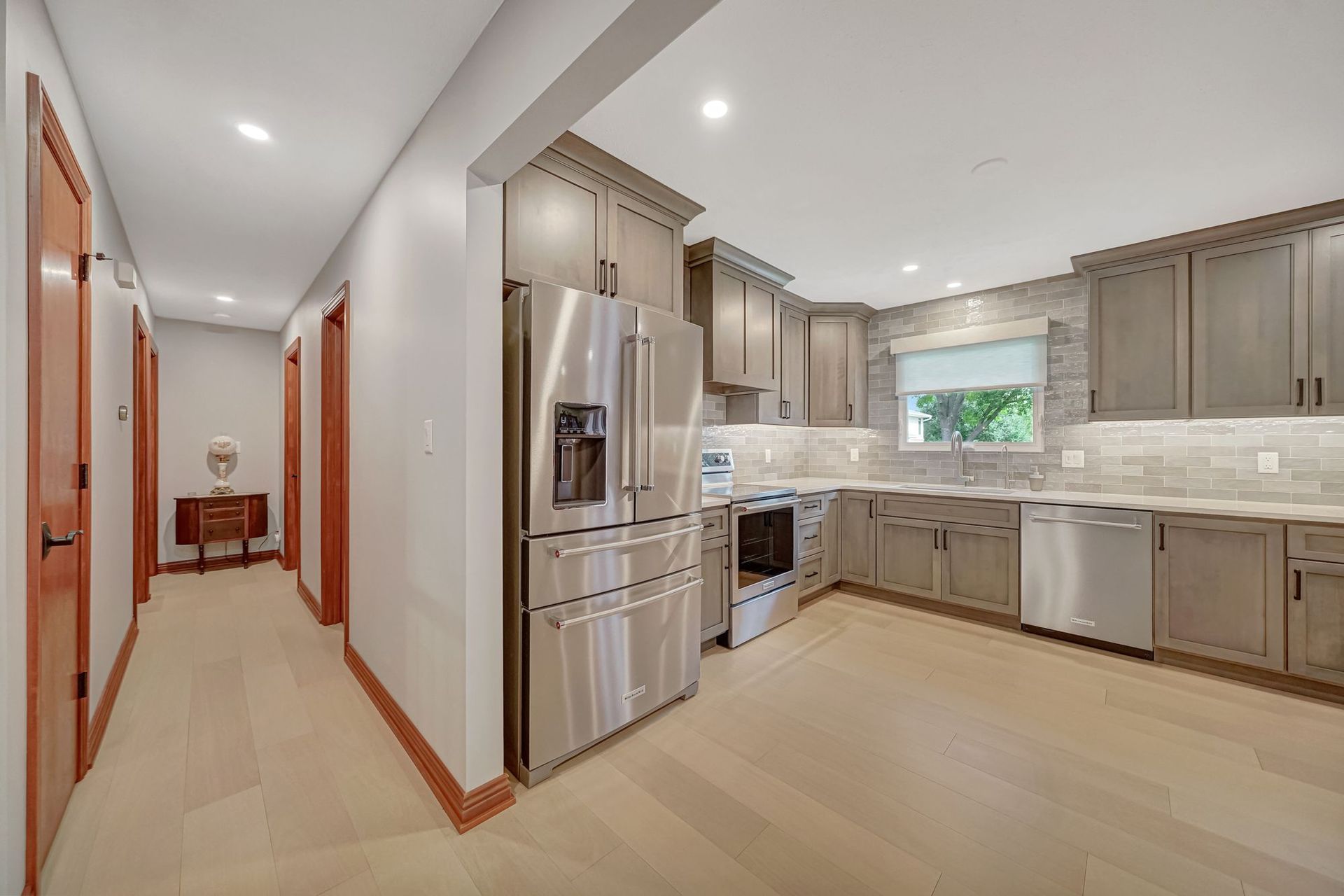 Kitchen with stainless steel appliances, gray cabinets, light wood floor, and hallway leading to doors.