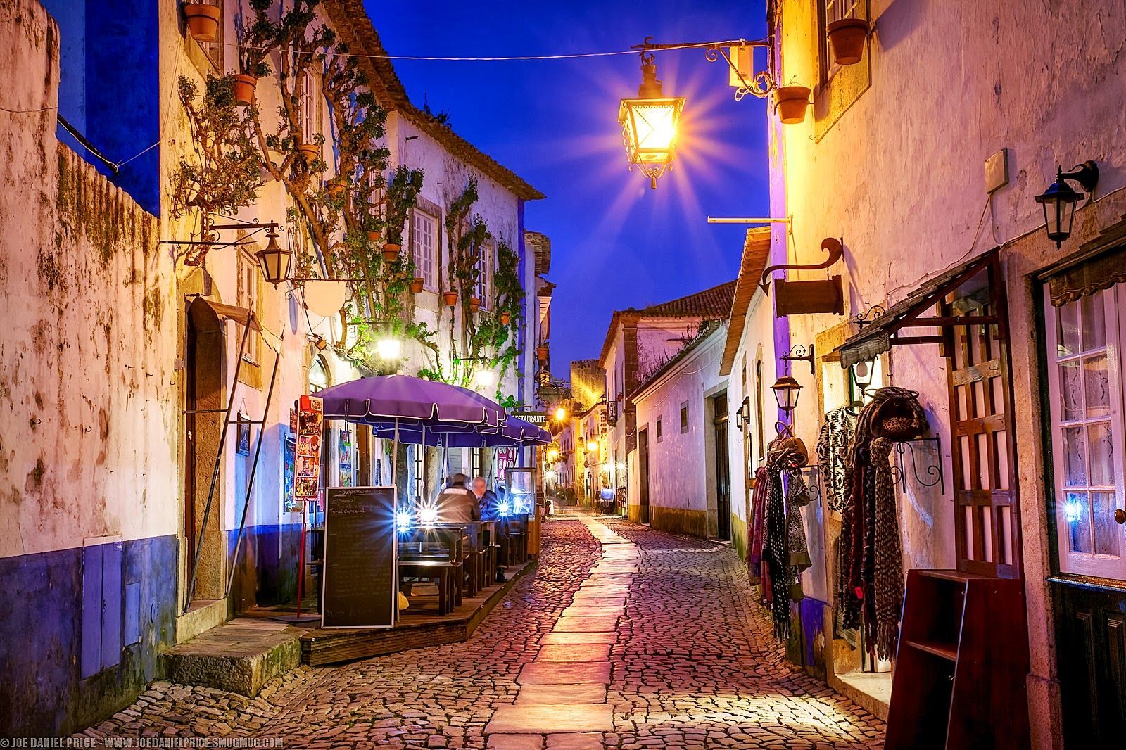 A narrow cobblestone street lined with buildings and shops at night.