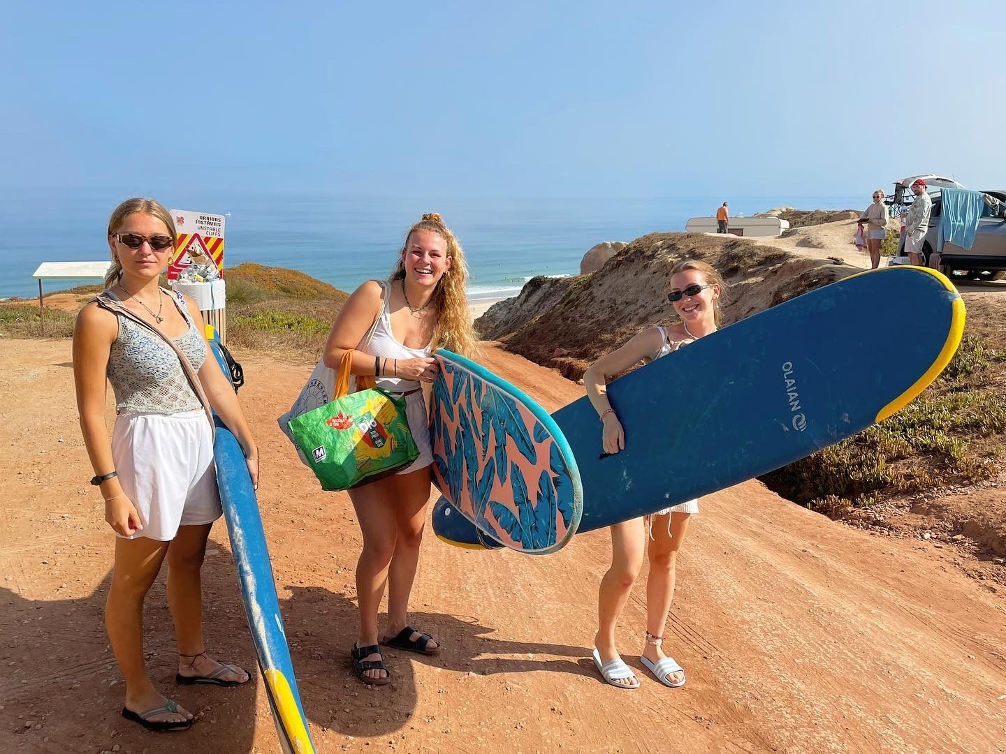 Three women are standing on a dirt road holding surfboards