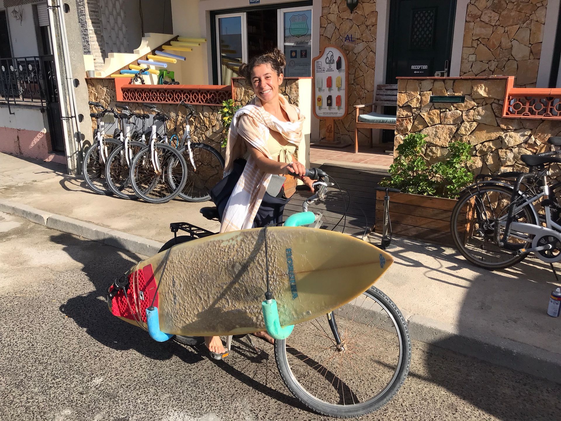 A woman is carrying a surfboard on a bike.
