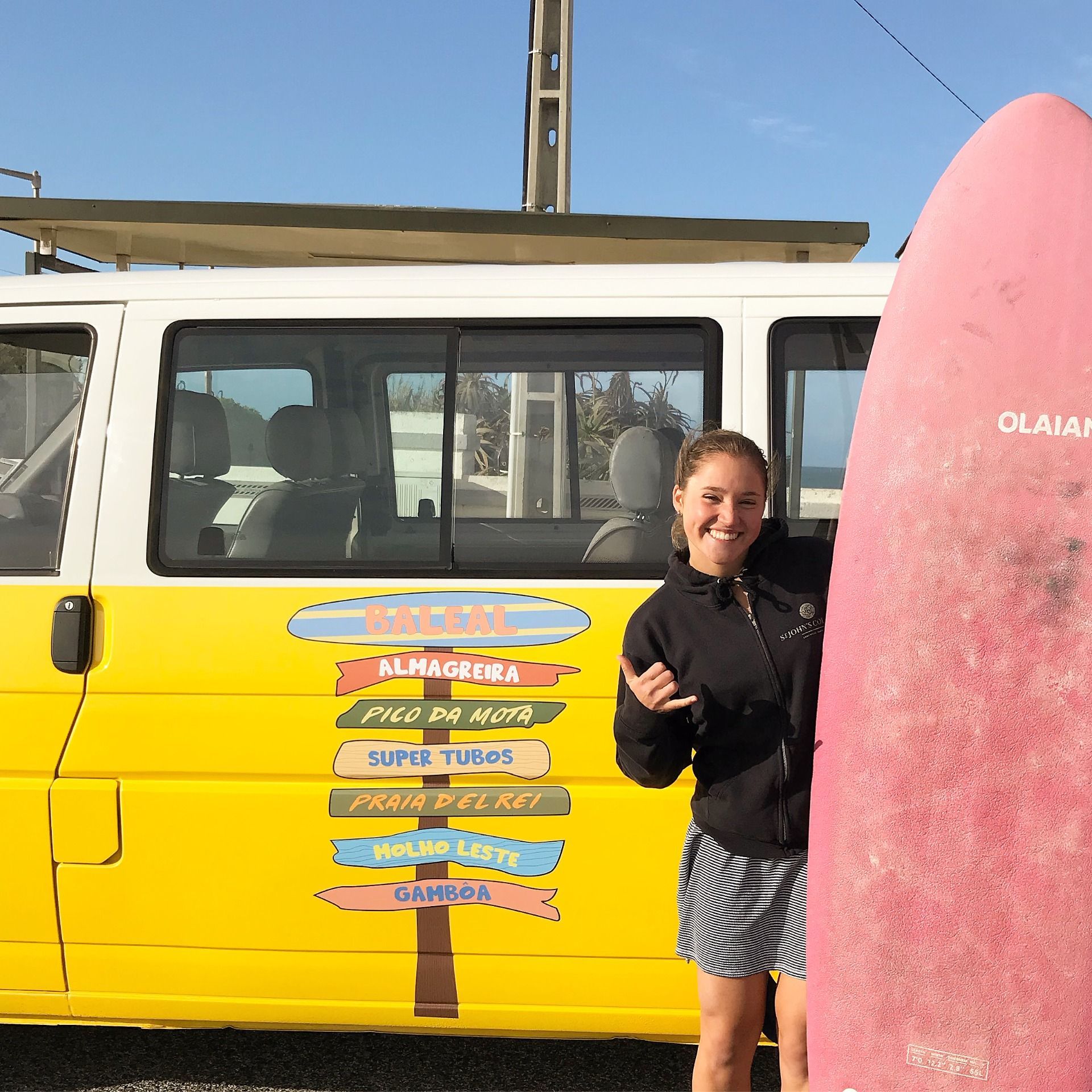 A woman stands next to a pink surfboard in front of a yellow van