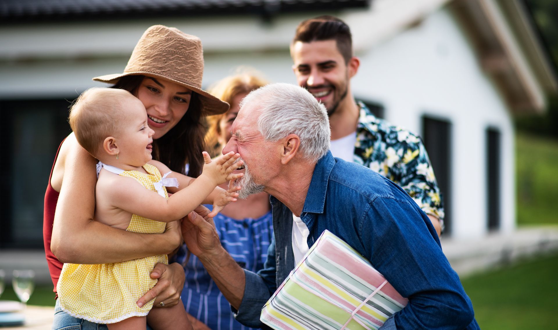 Family gathering outdoors; baby reaches toward elderly man, who holds a gift.