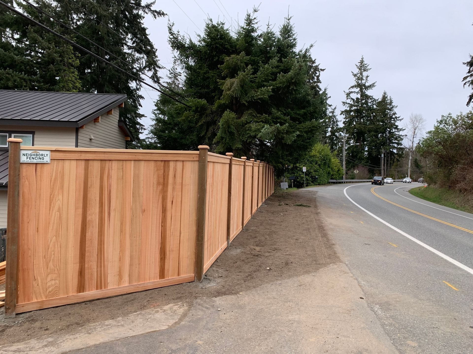 Wooden fence alongside a road.  Tall trees and a house in the background. Overcast sky.