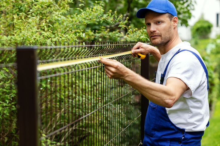 Man in blue overalls and cap measures a brown metal fence with a yellow tape measure, outdoors.