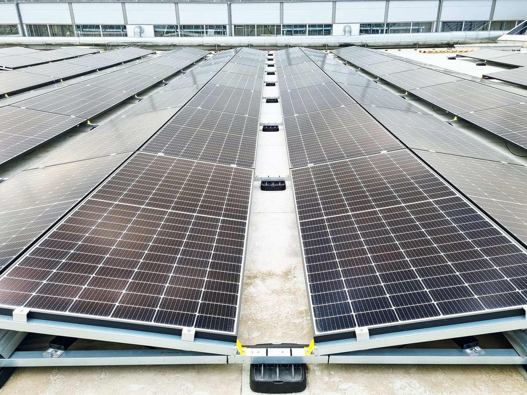 Rows of solar panels on a rooftop, viewed from the center aisle under bright daylight.