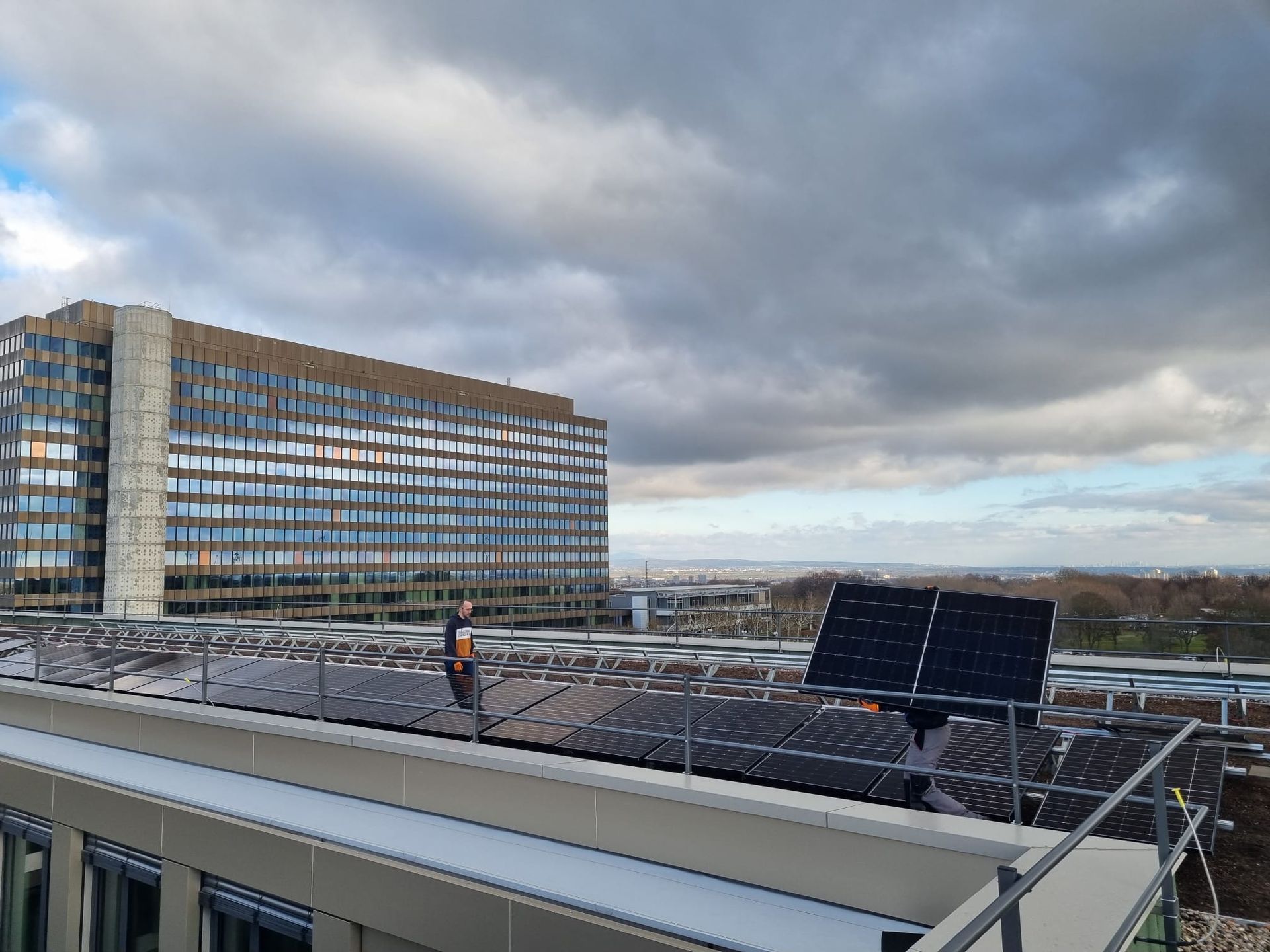 Rooftop solar panels with a person standing nearby, against a cloudy sky and office building.