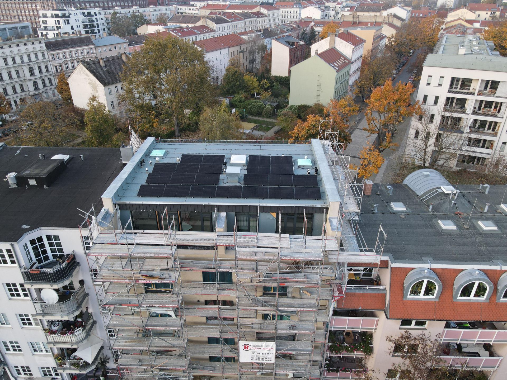 Aerial view of a building under renovation, covered in scaffolding amid surrounding city blocks and autumn trees.