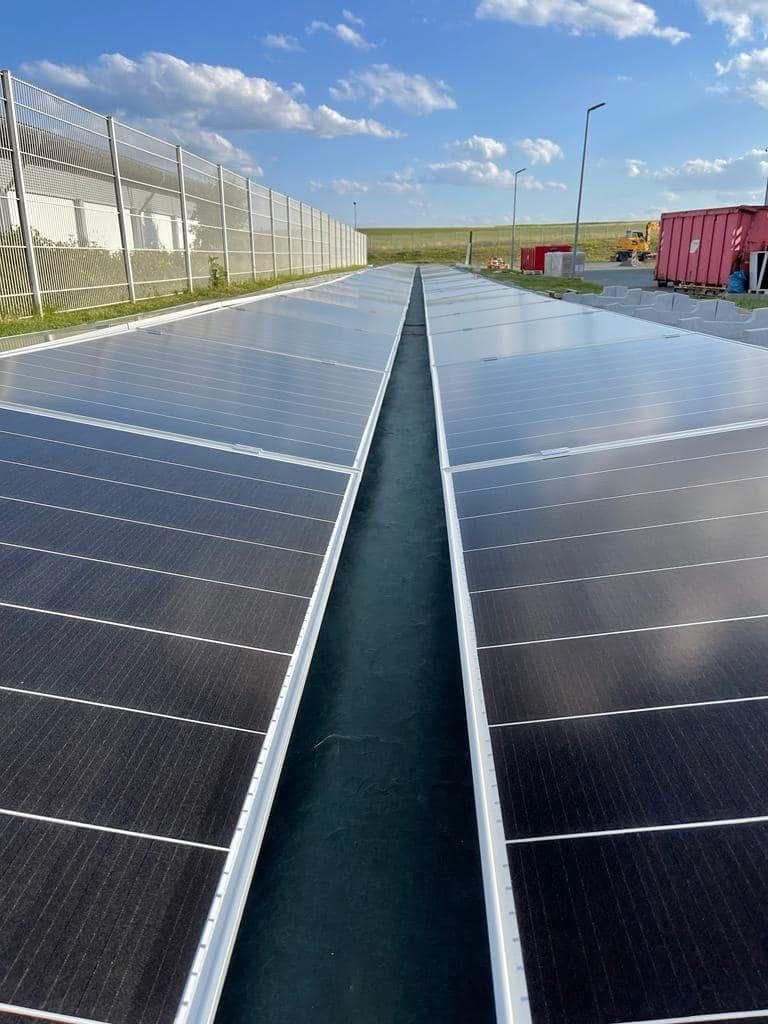 Rows of solar panels on a rooftop under a bright blue sky, with a fenced industrial area nearby