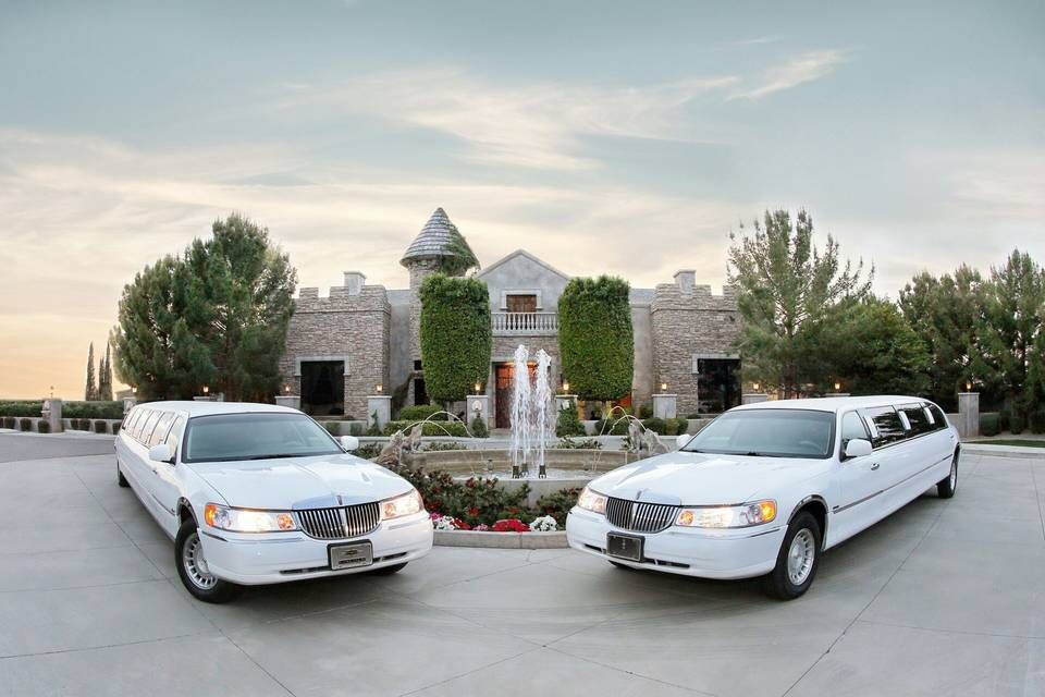 Two white limousines parked in front of a stone castle with a fountain.