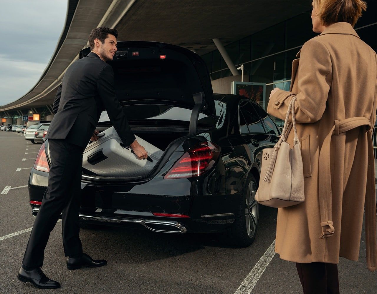 Man in suit loading luggage into the trunk of a black car at an airport; woman in coat stands nearby.