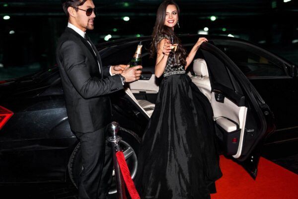 Man in suit pouring champagne as woman in formal dress exits a black car on a red carpet.