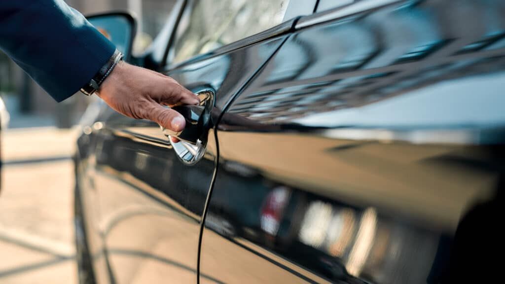 A person in a suit opening a black car door, likely in a city.