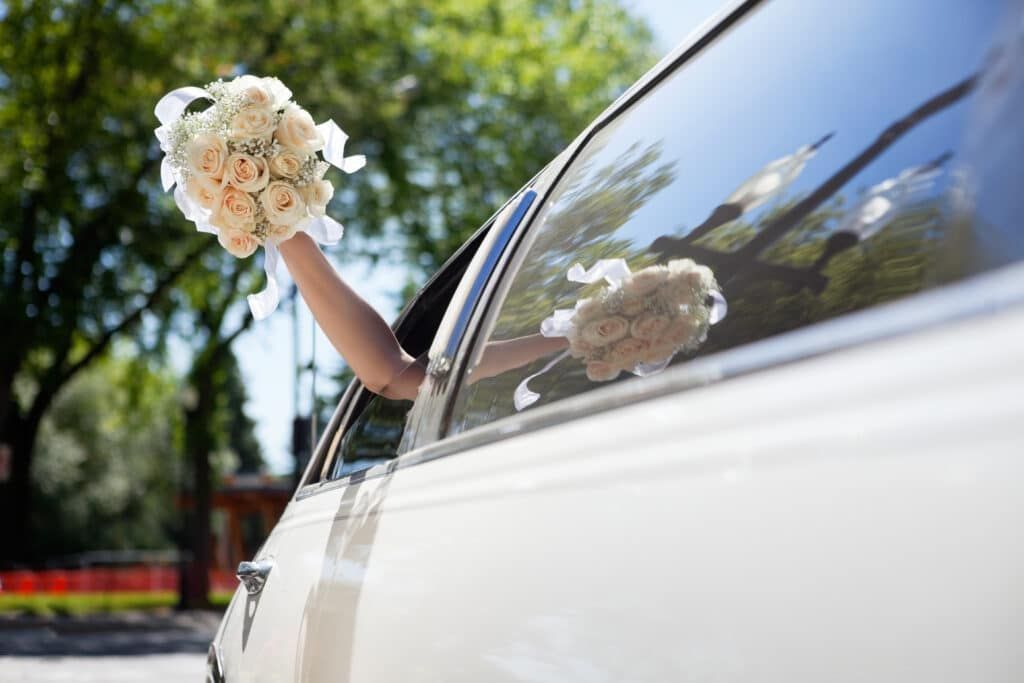 Bride waving a bouquet of white flowers from a car window on a sunny day.