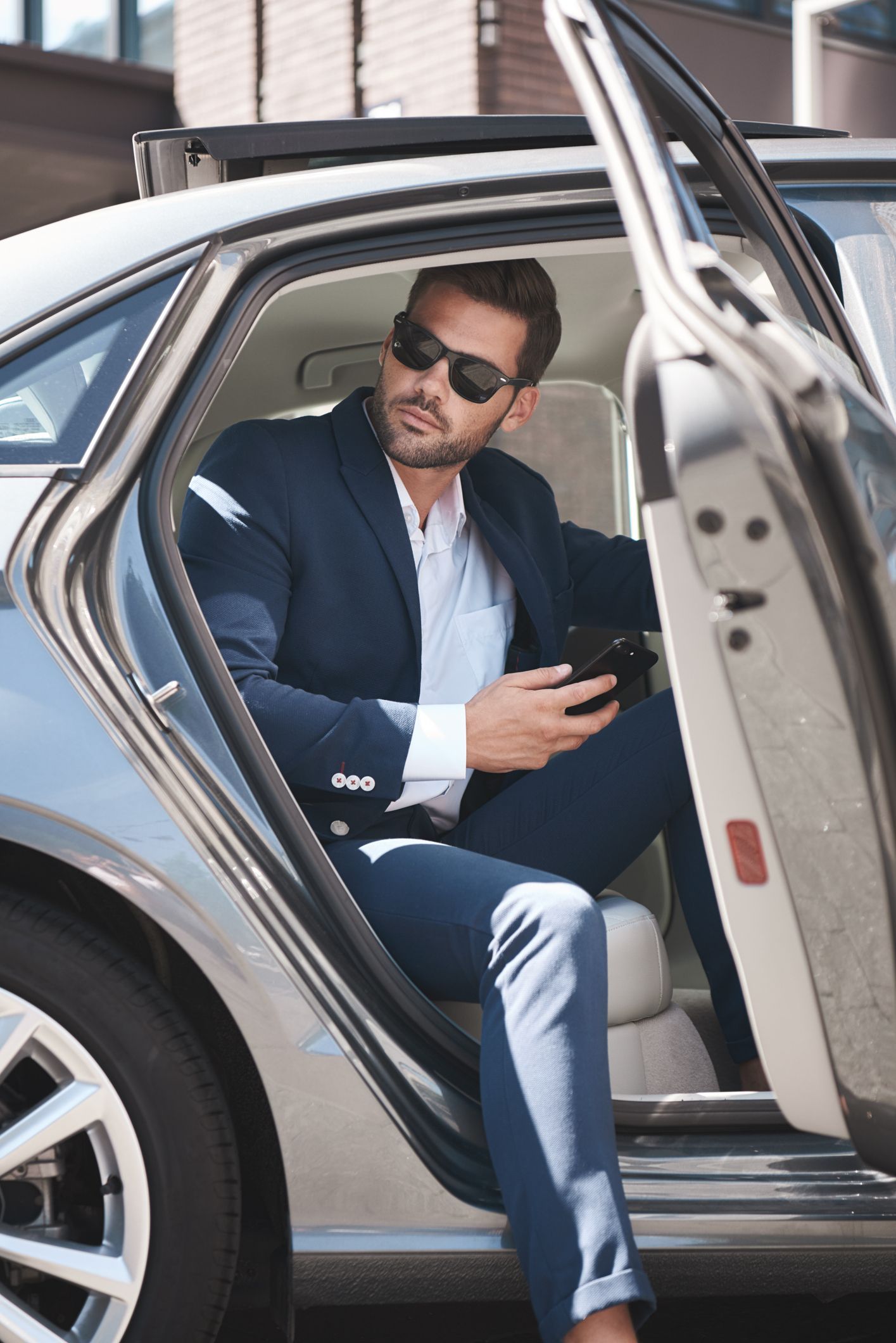 Man in suit and sunglasses exiting a car, holding a phone.