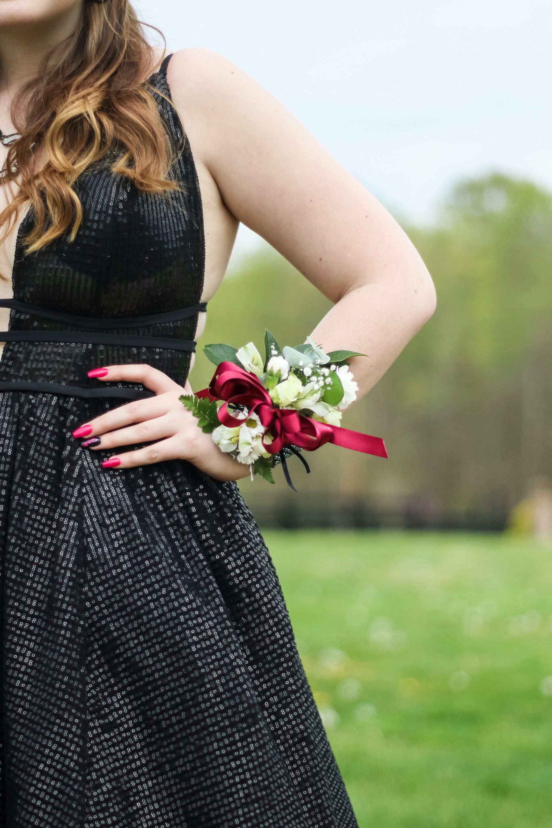 Woman in black sparkly dress with a corsage, hand on hip, outdoors in front of green foliage.