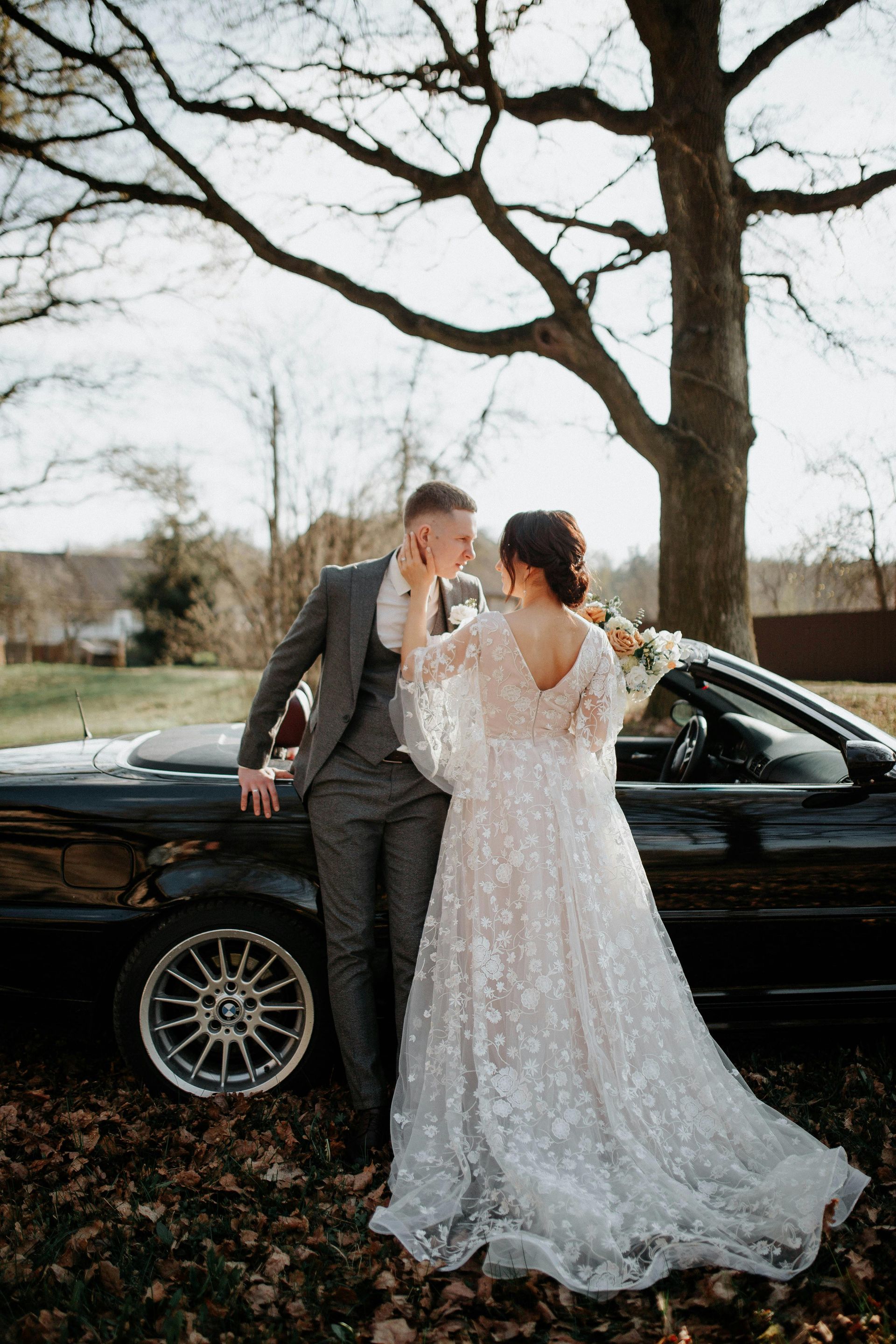 Bride and groom by a black convertible car, under a tree. The groom is in a gray suit, and the bride wears a sparkling white 