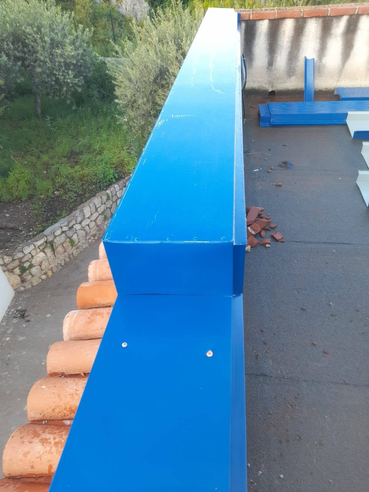 Grondaia o canale di scolo in metallo blu sul tetto, accanto a tegole arancioni e un sentiero di ghiaia all'esterno.