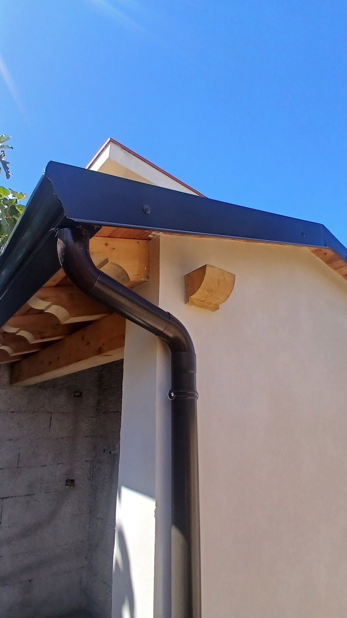 Esterno del tetto di una casa con grondaia nera e muro beige sotto un cielo azzurro brillante