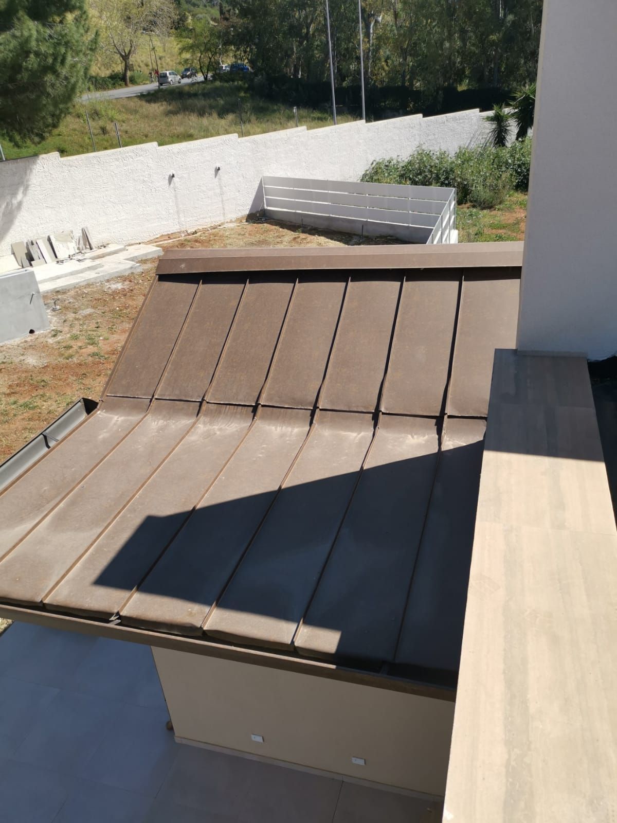 Terrazza esterna con tetto in metallo inclinato marrone accanto a una parete bianca e alberi da giardino sullo sfondo.
