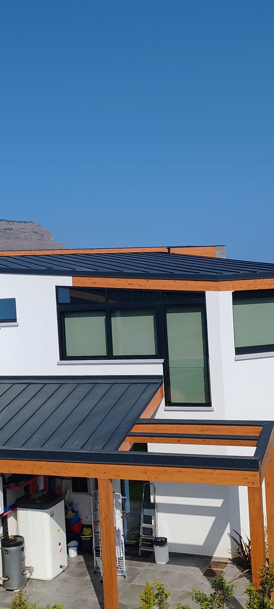 Casa moderna a due piani con tetto grigio-blu, pareti bianche, ampie finestre e un portico coperto sotto un cielo azzurro limpido.