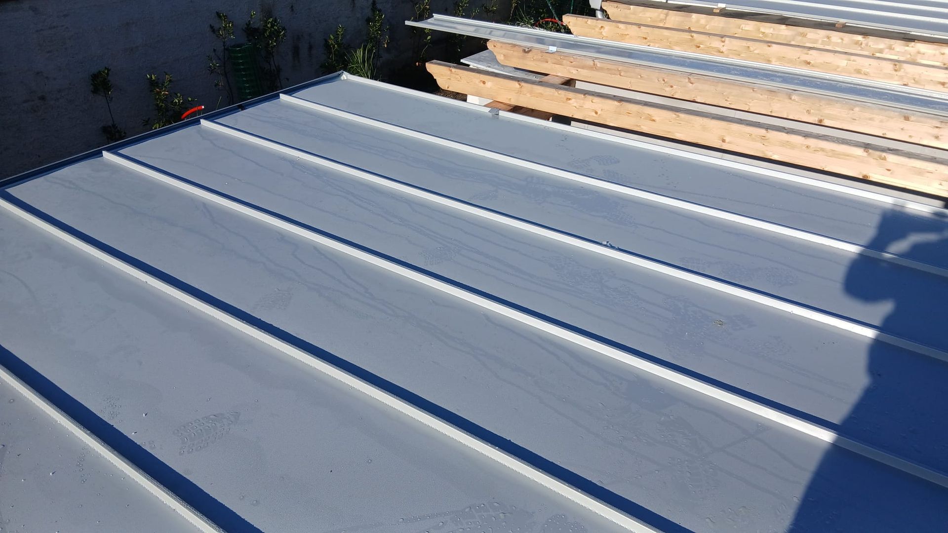 Pannelli di metallo bianco per il tetto accanto a una struttura in legno a vista illuminata dalla luce del sole, con l'ombra di una persona sulla destra.
